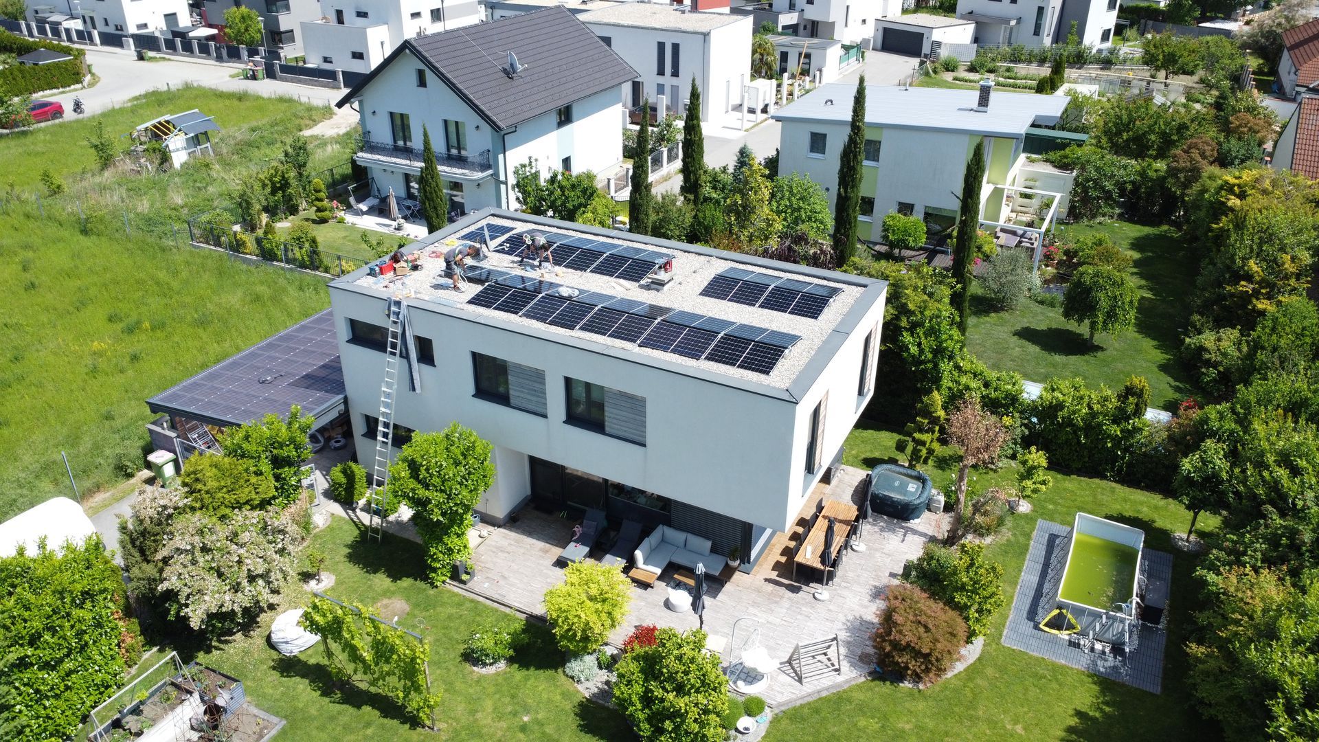 Luftaufnahme eines weißen Hauses mit Solarpaneelen, umgeben von einem grünen Garten und Nachbarhäusern.