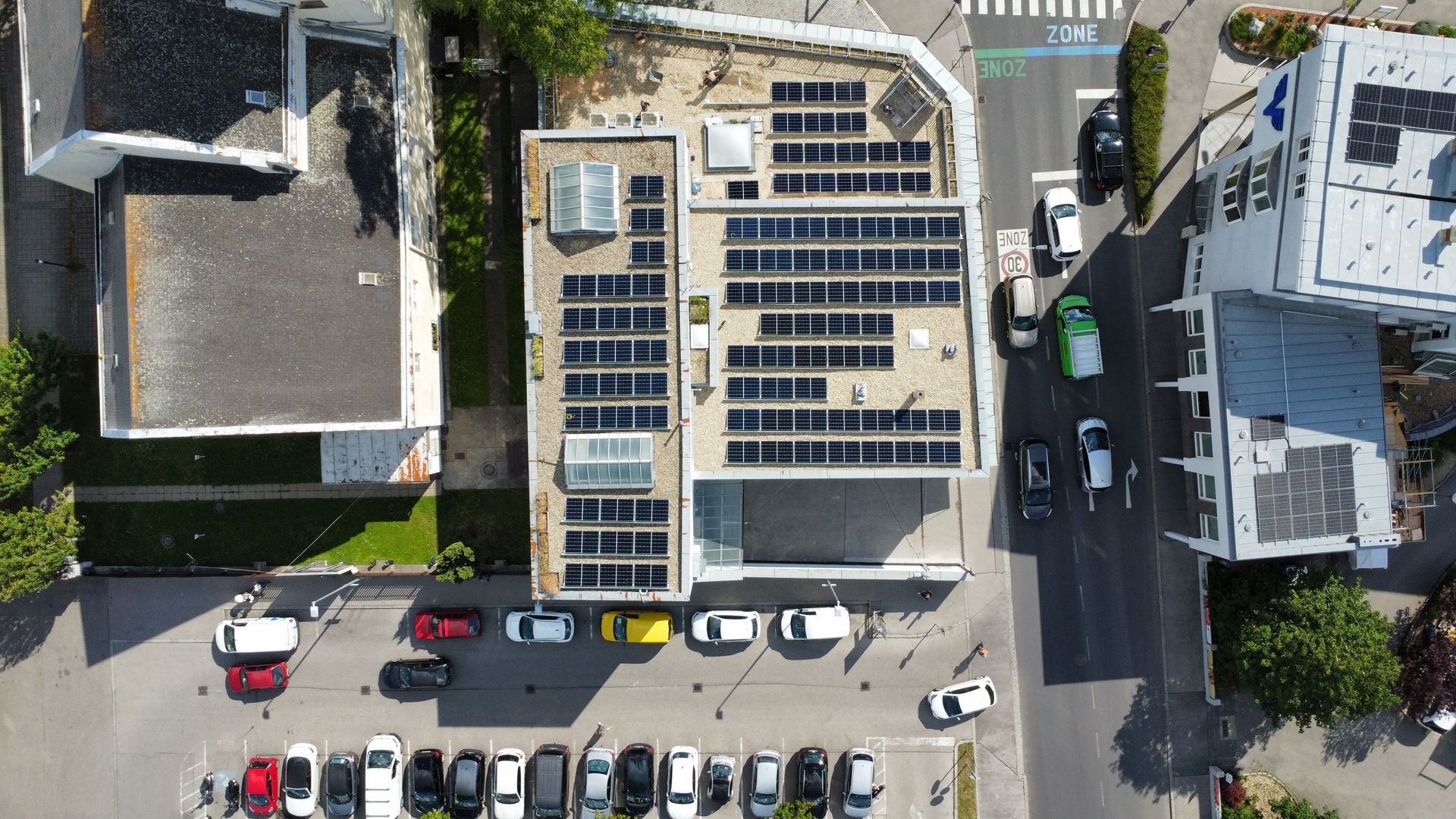 Luftaufnahme eines mit Solarpaneelen bedeckten Gebäudedachs neben geparkten Autos auf einer Straße.