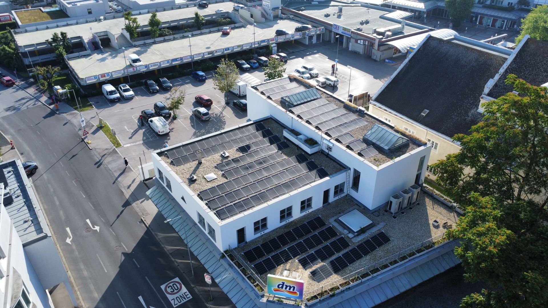 Luftaufnahme eines weißen Gebäudes mit Flachdach neben einer Straße und einem Parkplatz, mit Bäumen und umliegenden Gebäuden.