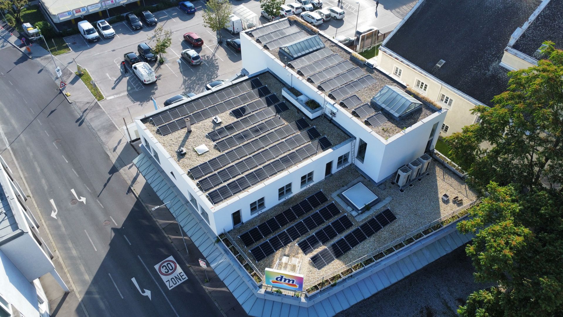 Luftaufnahme eines weißen Gebäudes mit Solarpaneelen auf dem Dach neben einer Straße und Bäumen.
