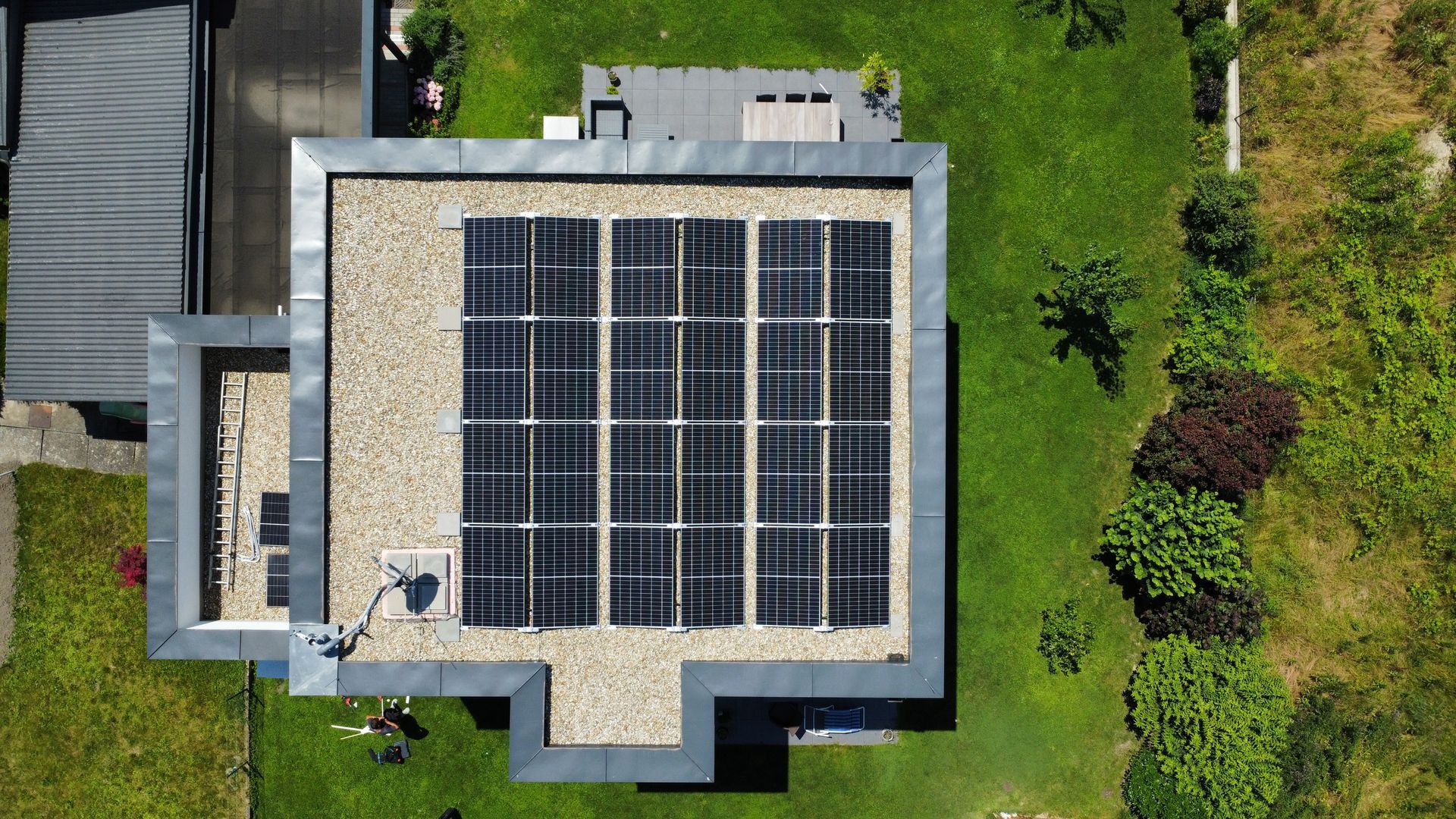 Luftaufnahme eines Flachdachs mit Solarpaneelen, umgeben von grünen Bäumen und einem Garten.