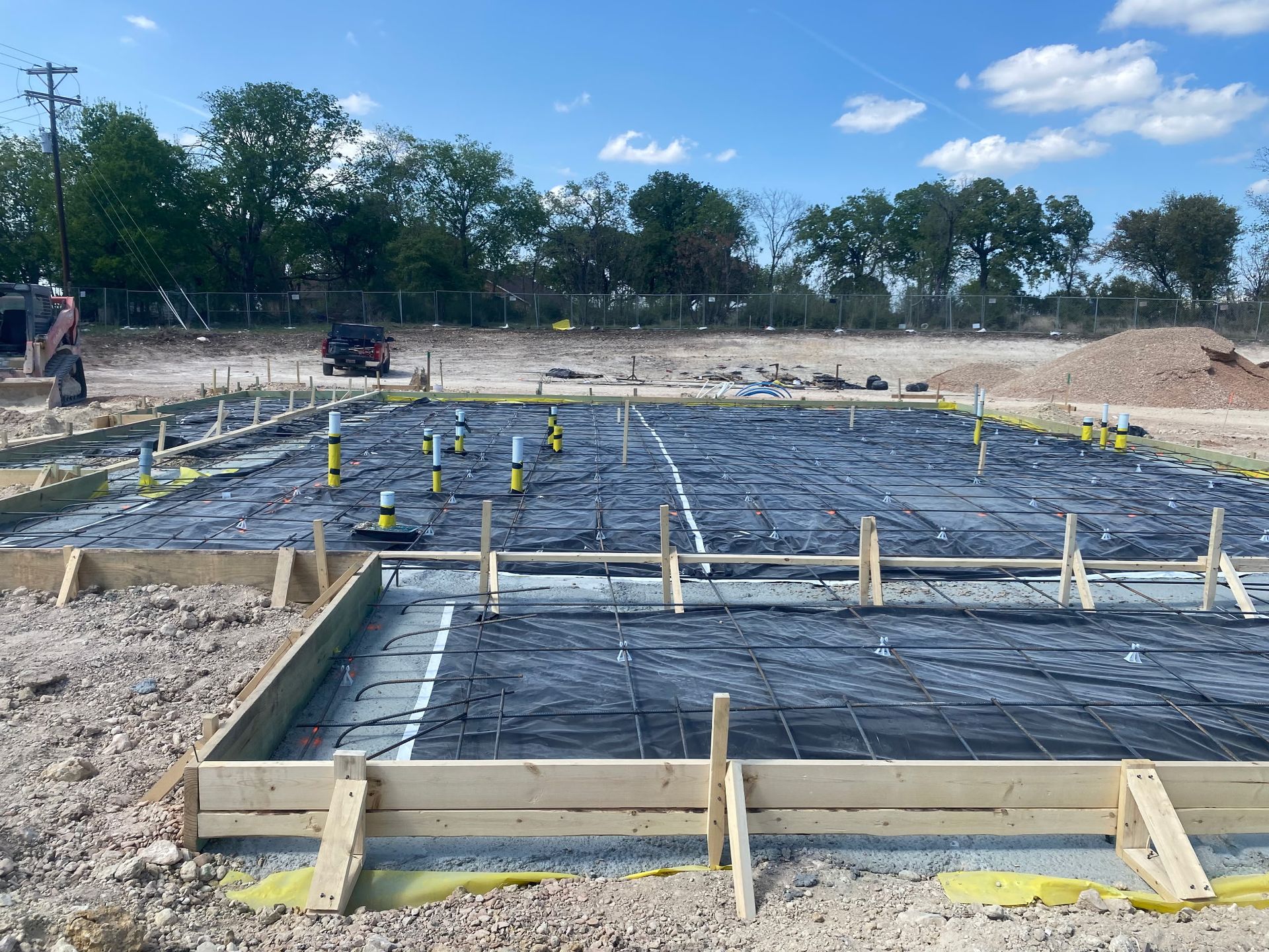 Foundation under construction with rebar grid, wooden forms, and yellow markers on a dirt site.