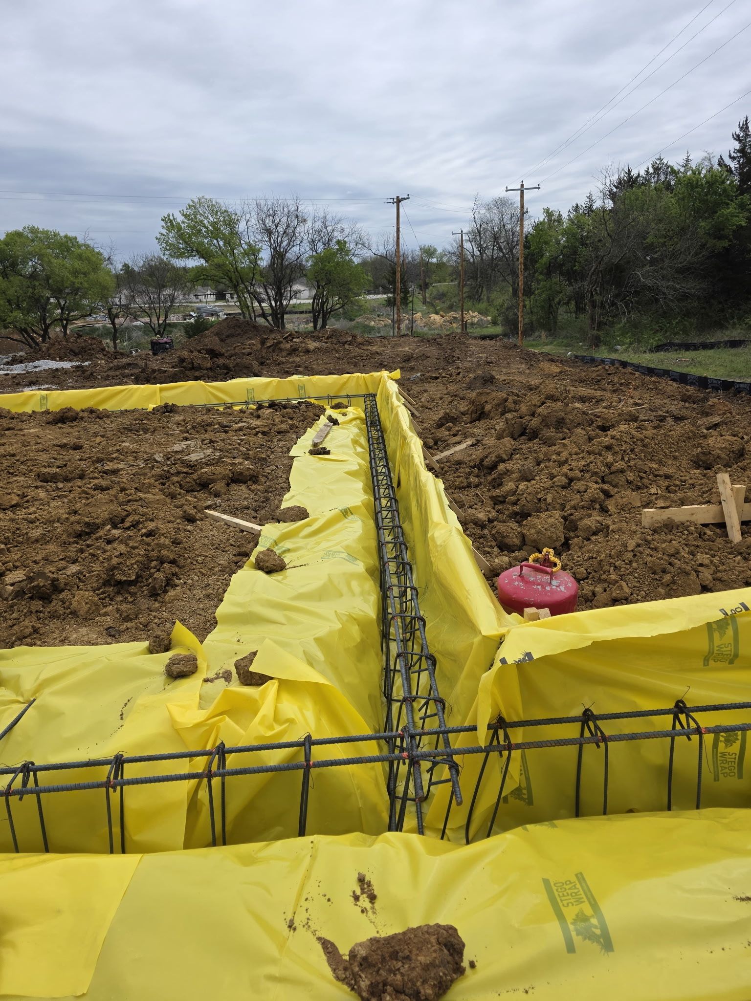 Yellow sheeting around muddy excavation site with rebar, dirt piles, and cloudy sky
