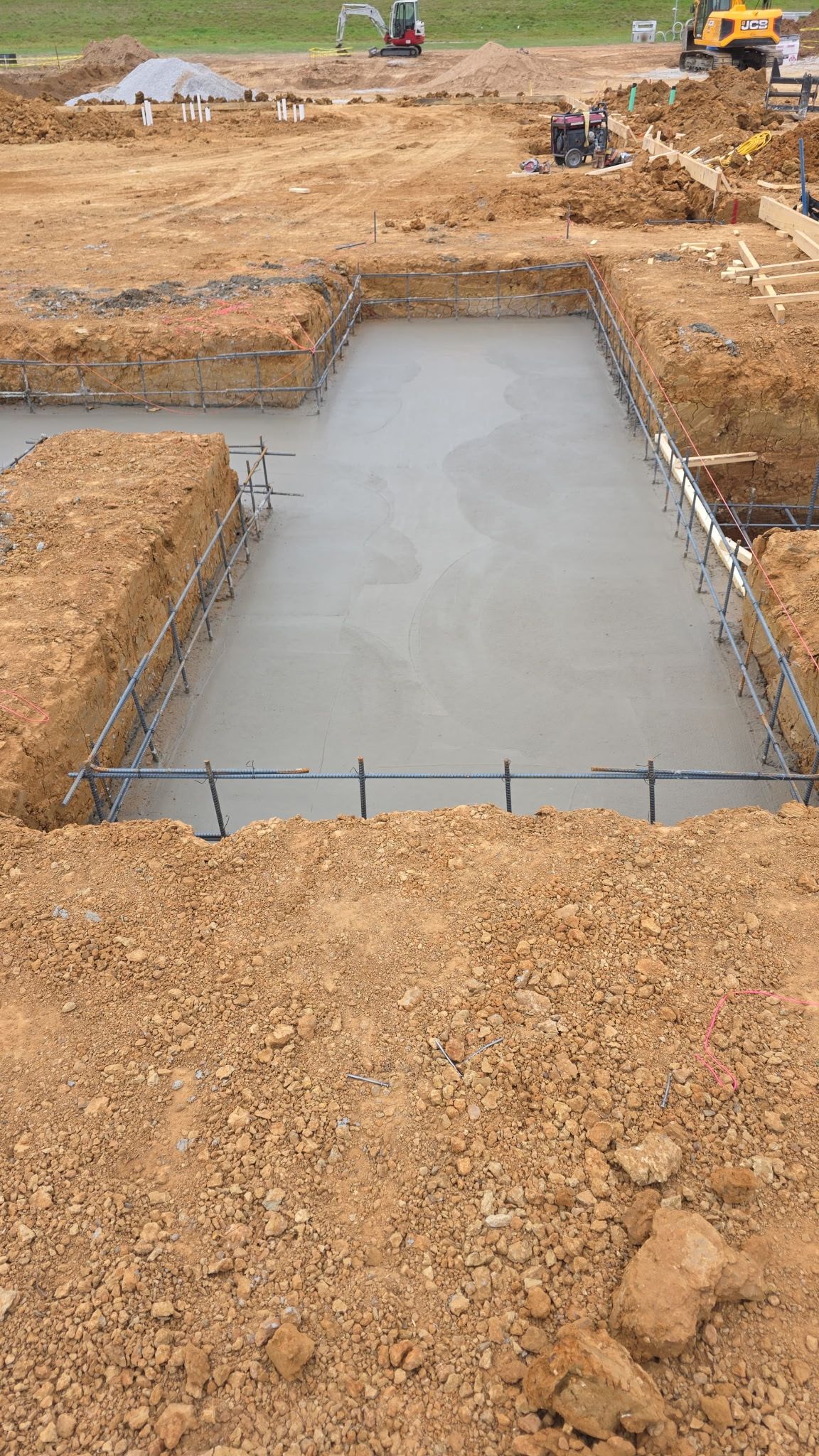 Fresh concrete foundation slab at a construction site, surrounded by dirt and gravel, with equipment in the background