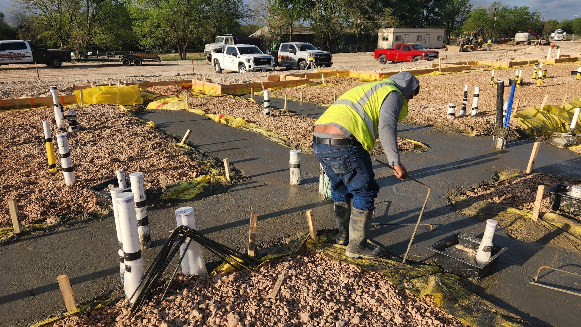 Worker in a safety vest and red hard hat pouring concrete at a construction site near leaf-covered ground.