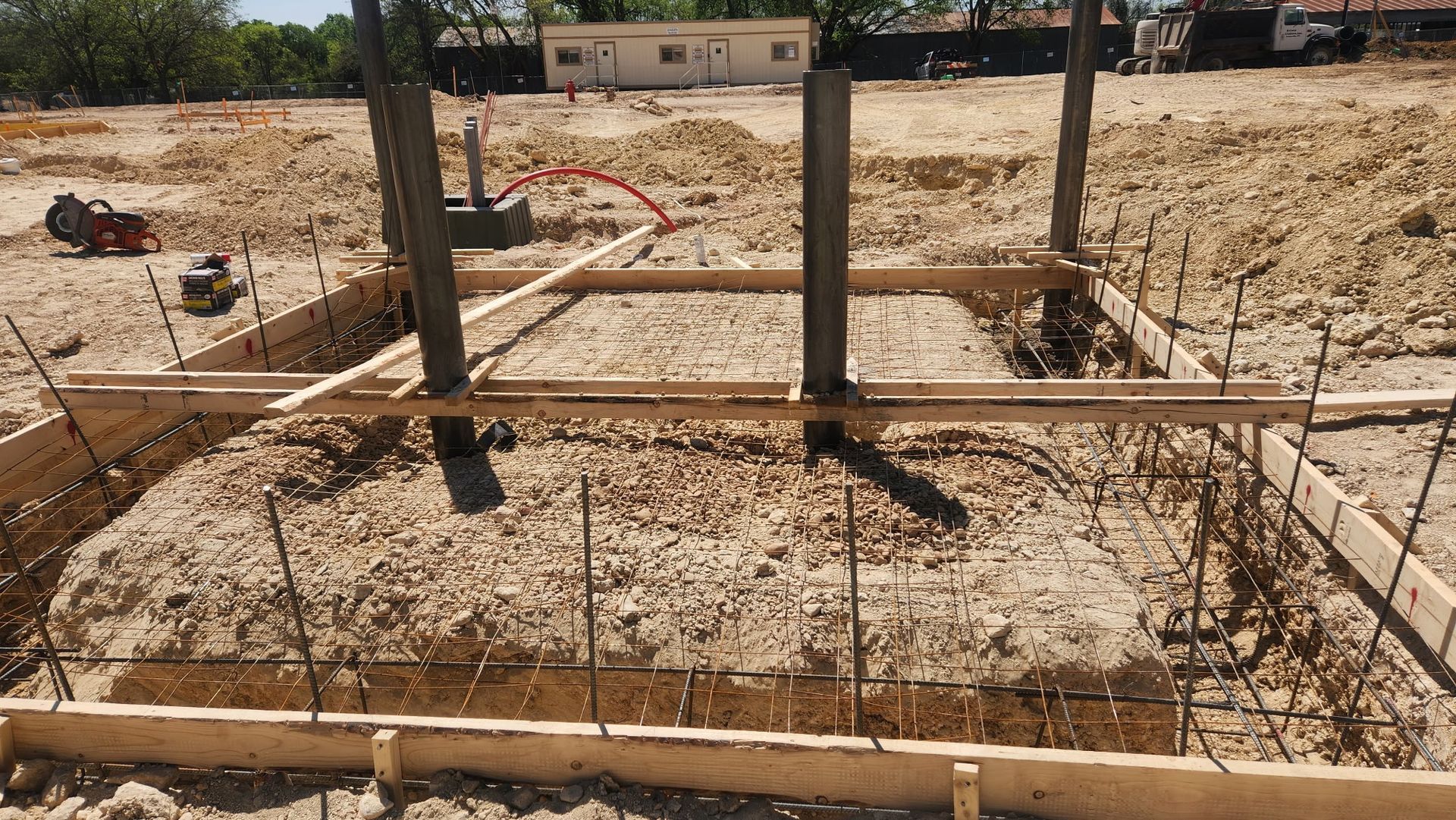 Construction site foundation with wooden forms, rebar, and two vertical steel columns on sandy ground