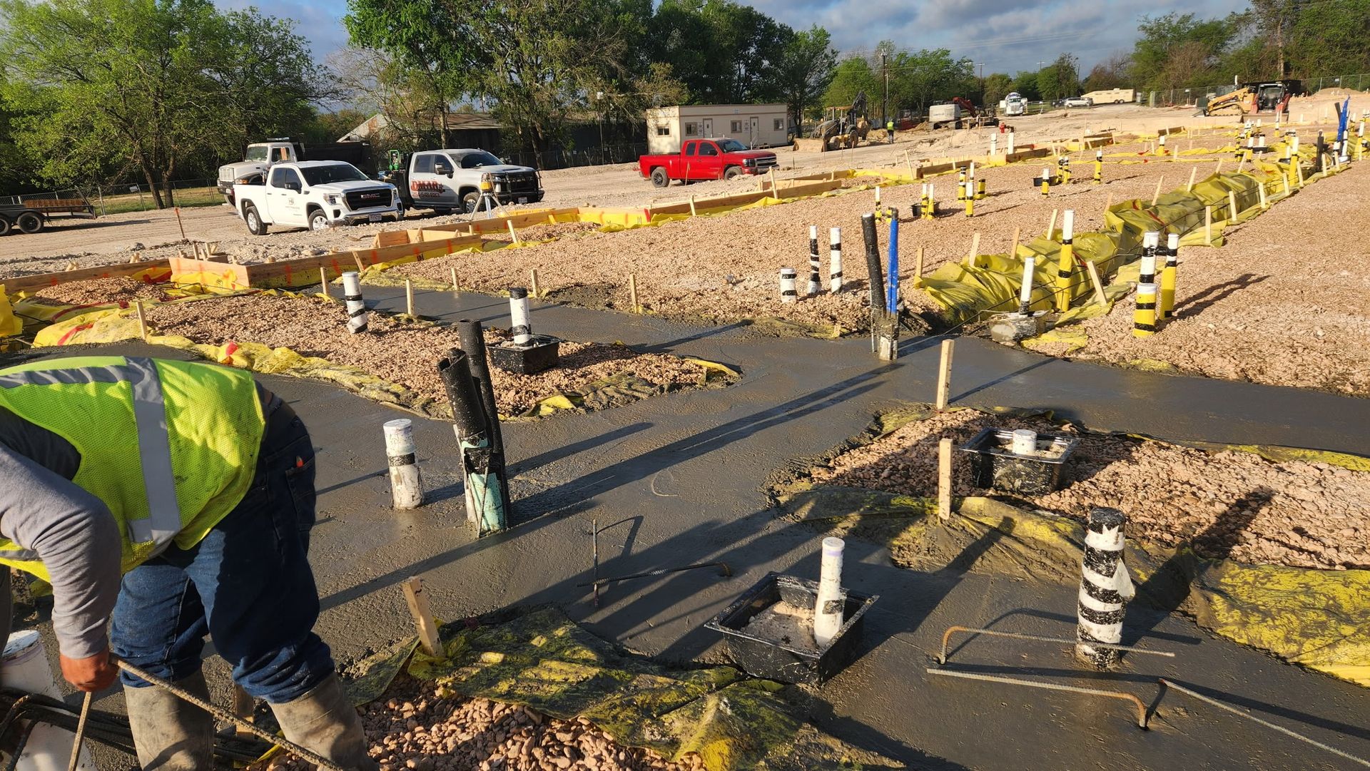 Roadwork site with workers laying dark pavement strips beside parked vehicles and safety cones.