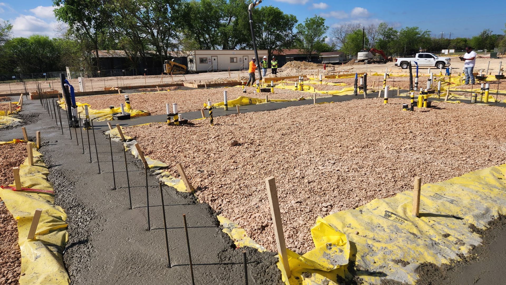 Construction site with a concrete foundation, rebar, yellow barriers, and gravel under a clear sky