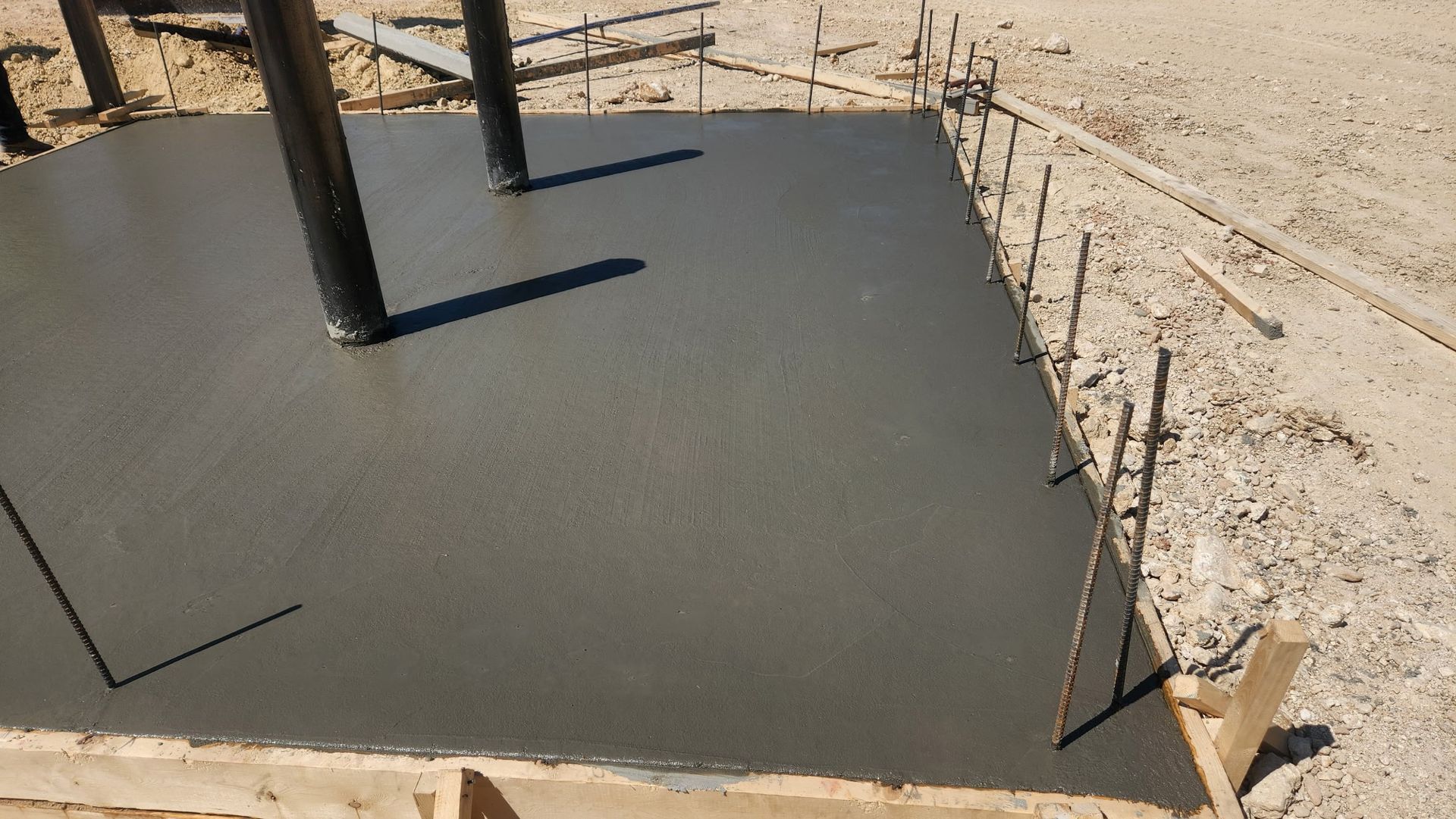 Fresh concrete slab foundation with black support posts at a sandy construction site