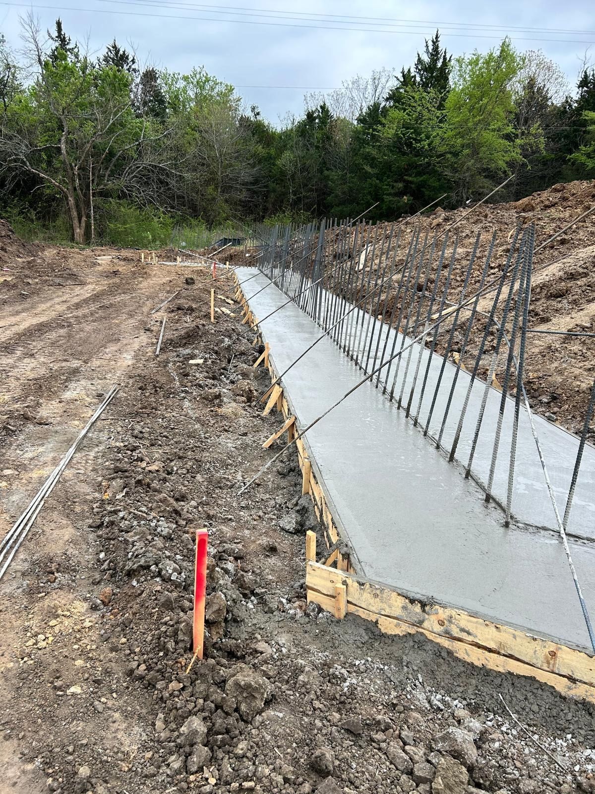 Fresh concrete foundation along a dirt trench, with rebar mesh and a red survey stake in a wooded area