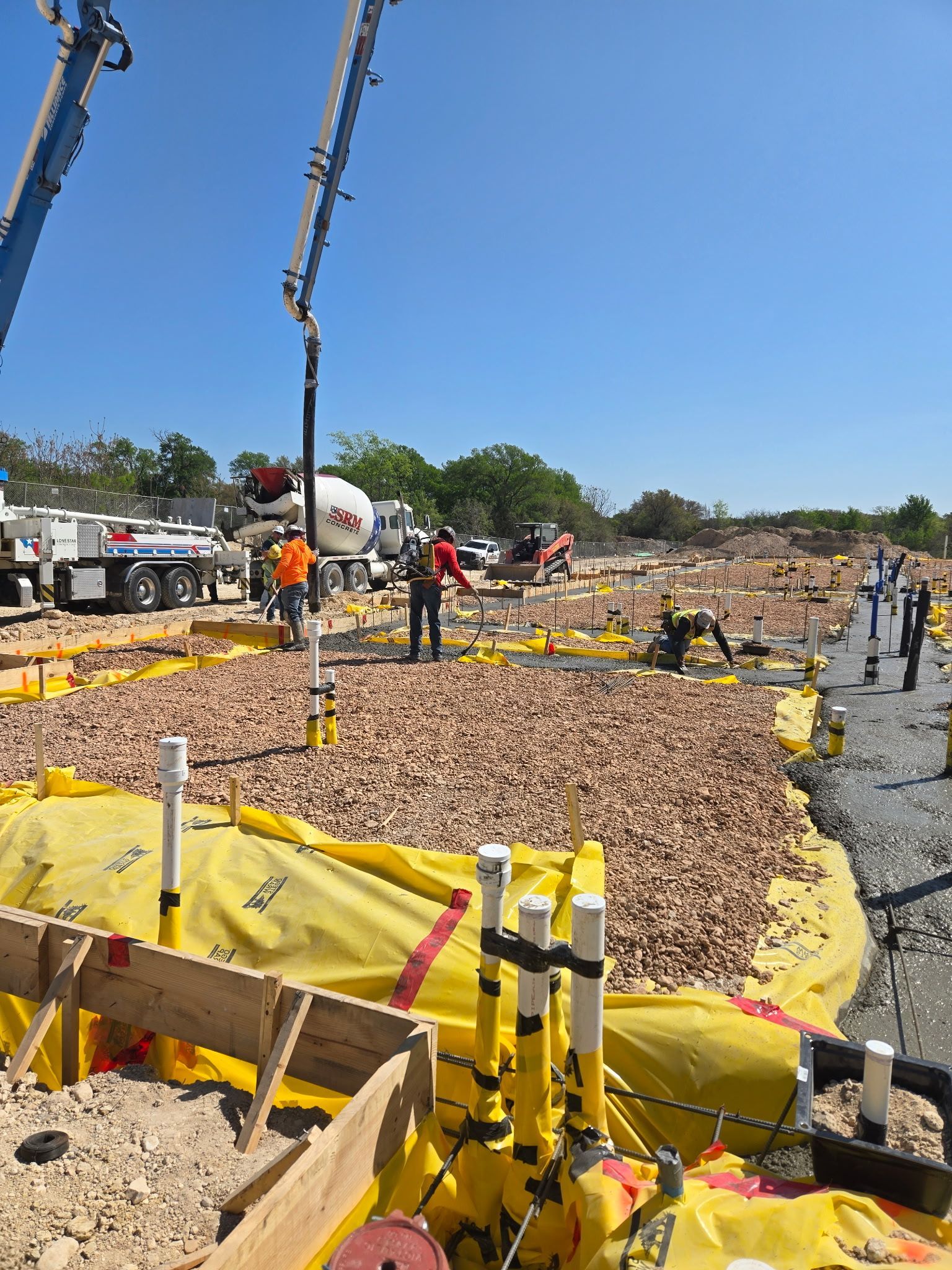 Construction site with concrete pump truck, workers, and yellow barriers over a rocky foundation