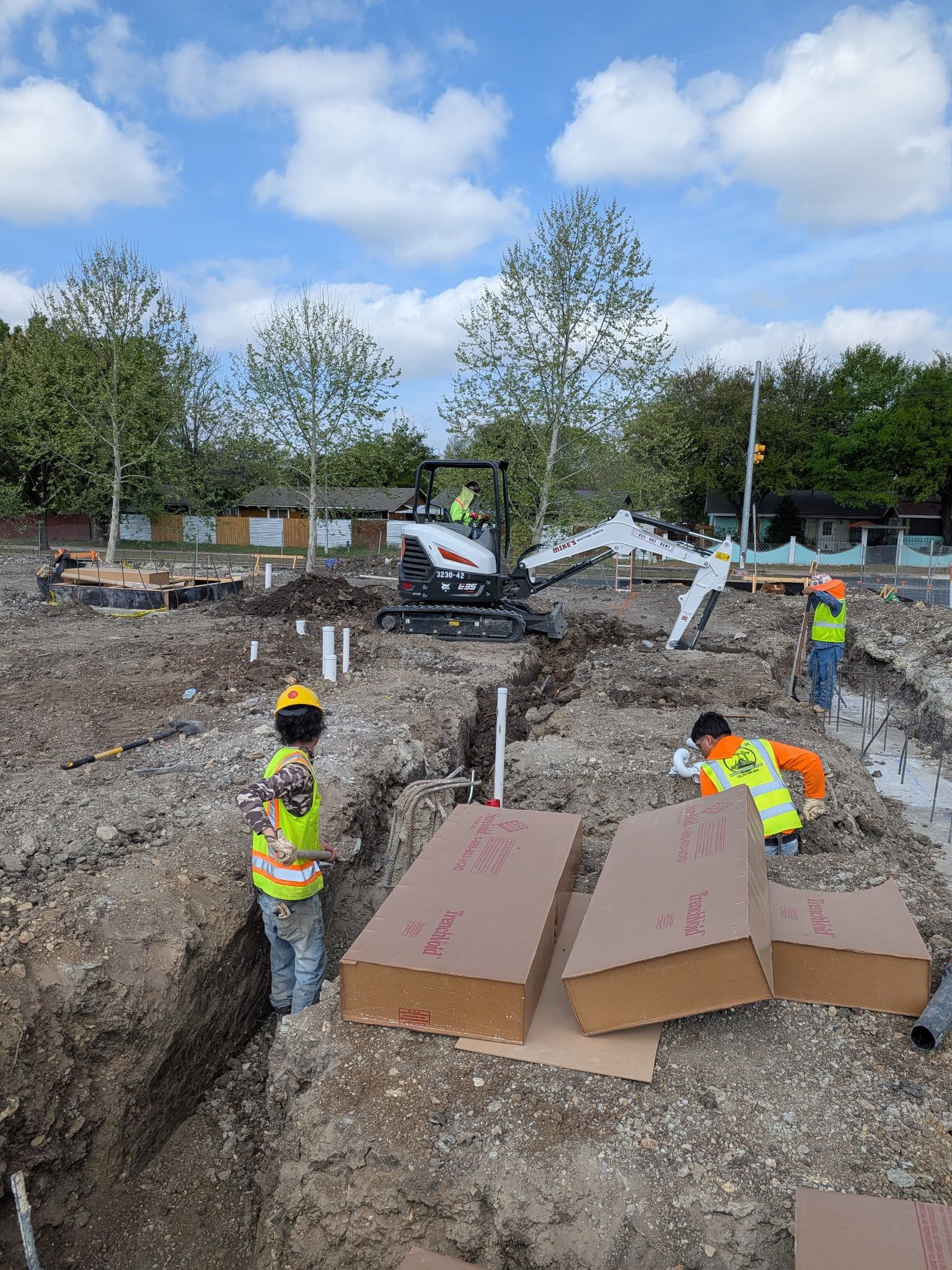 Workers in hard hats standing in a muddy construction site near large cardboard boxes and excavated earth.