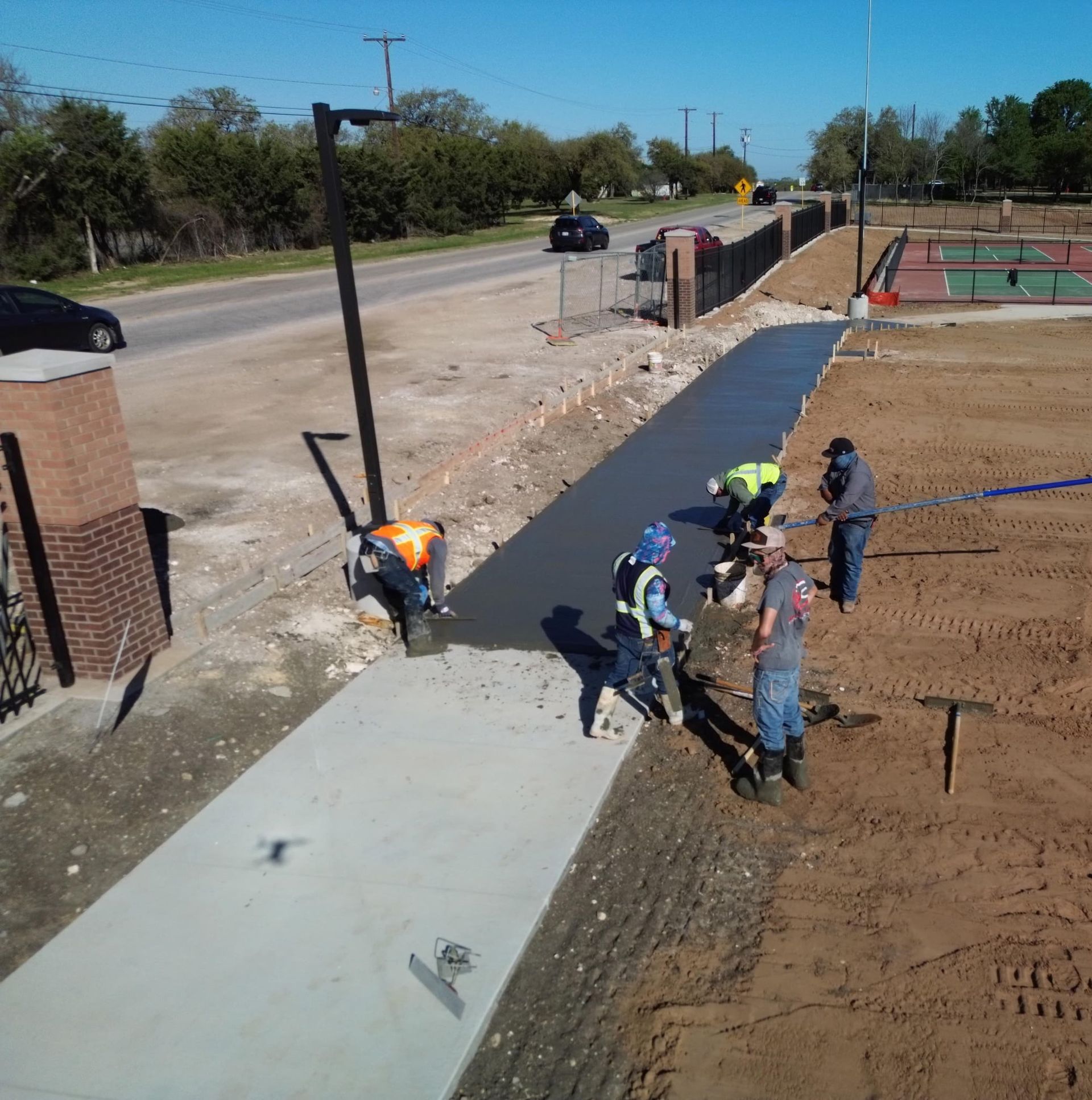 Workers pour concrete beside a road at a construction site near a sports field.