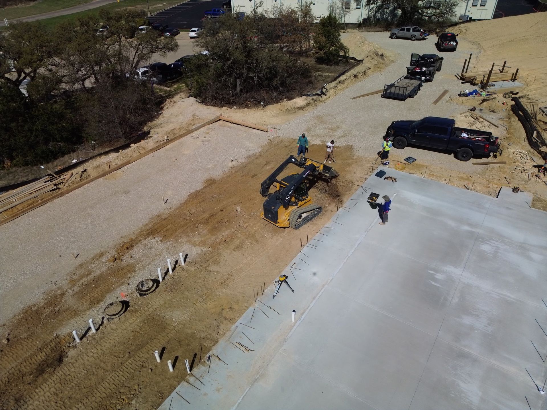 Roadwork scene with a yellow excavator beside a concrete slab and parked trucks on a dirt site.