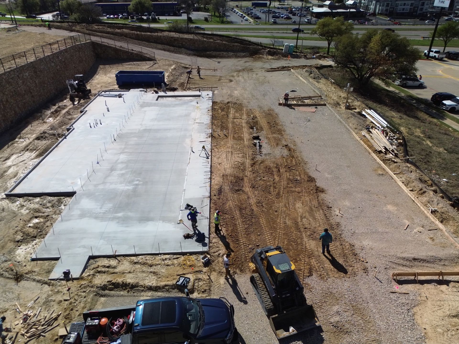 Construction site with concrete slab, dirt lot, and heavy equipment beside a road and retaining wall