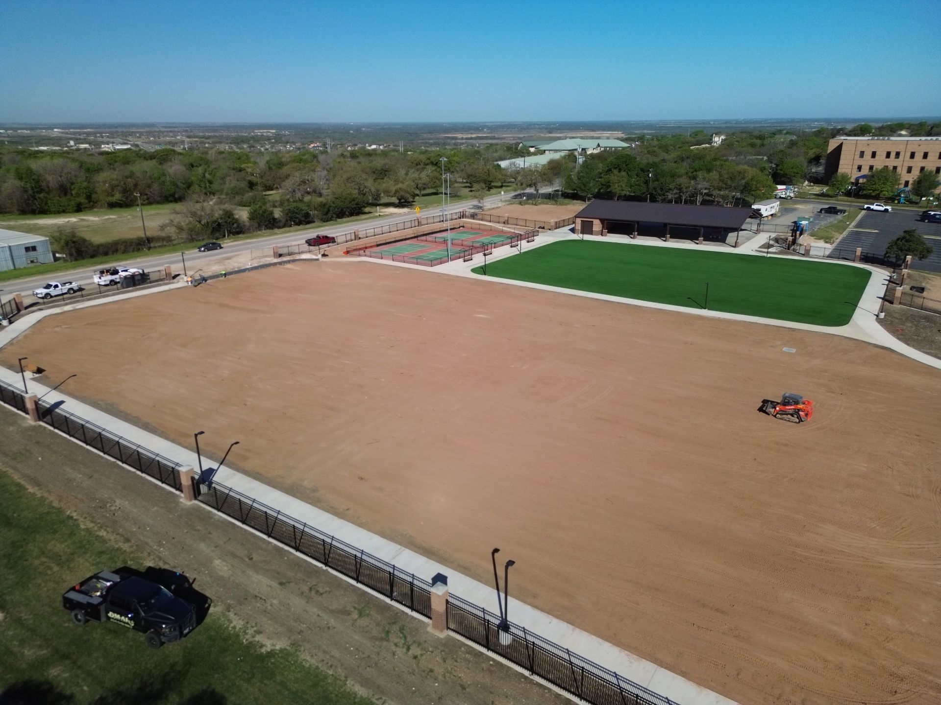 Aerial view of a large dirt field beside a green sports court and buildings, with a few people on the field.