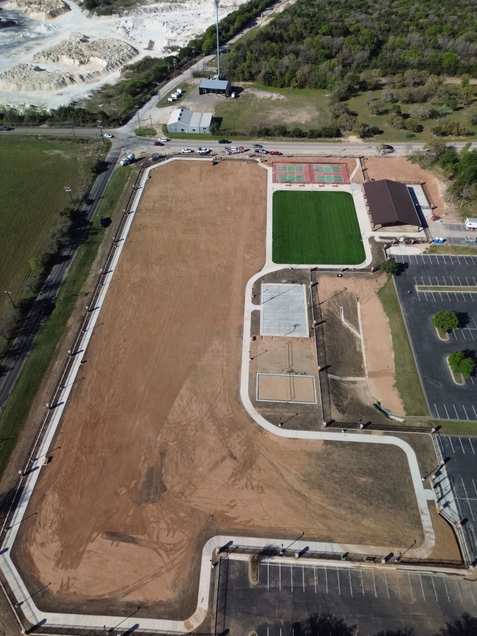 Aerial view of a sports field complex with a green turf field, brown dirt lot, and parking area.