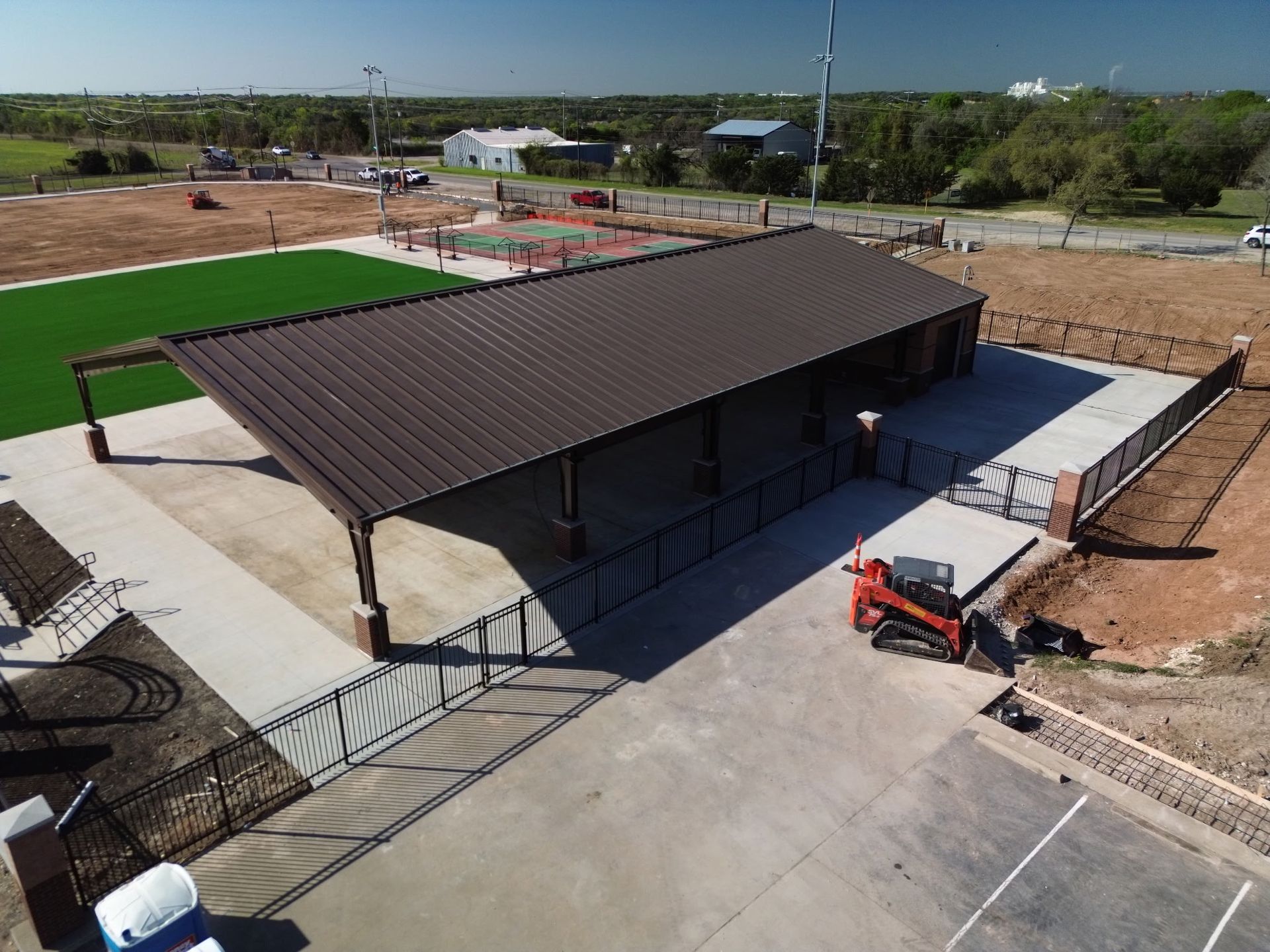 Aerial view of a pavilion with a brown roof, concrete plaza, and nearby construction equipment in a park area