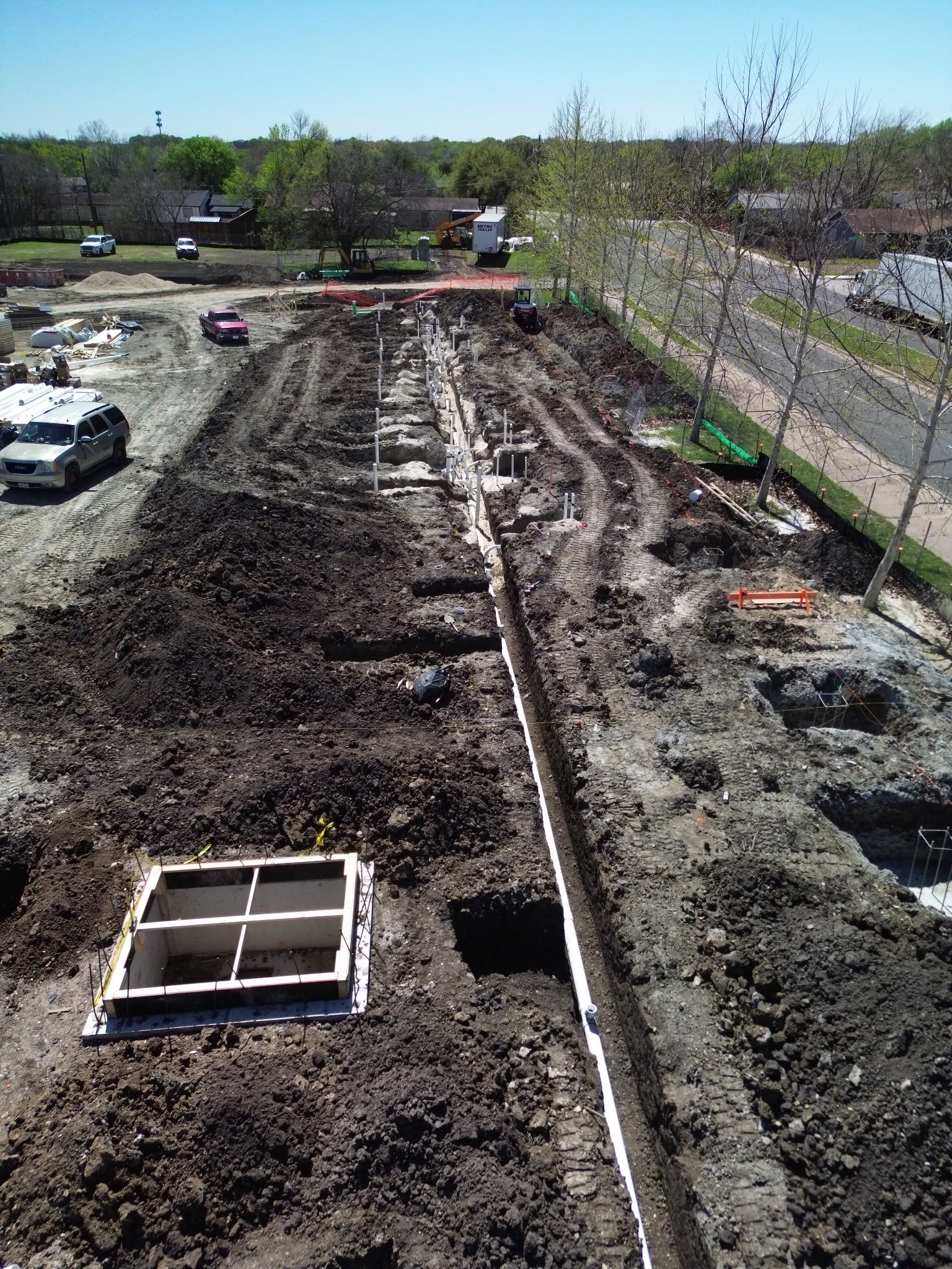 Construction site with a long muddy trench, exposed pipes, and a white vehicle at left under a clear sky