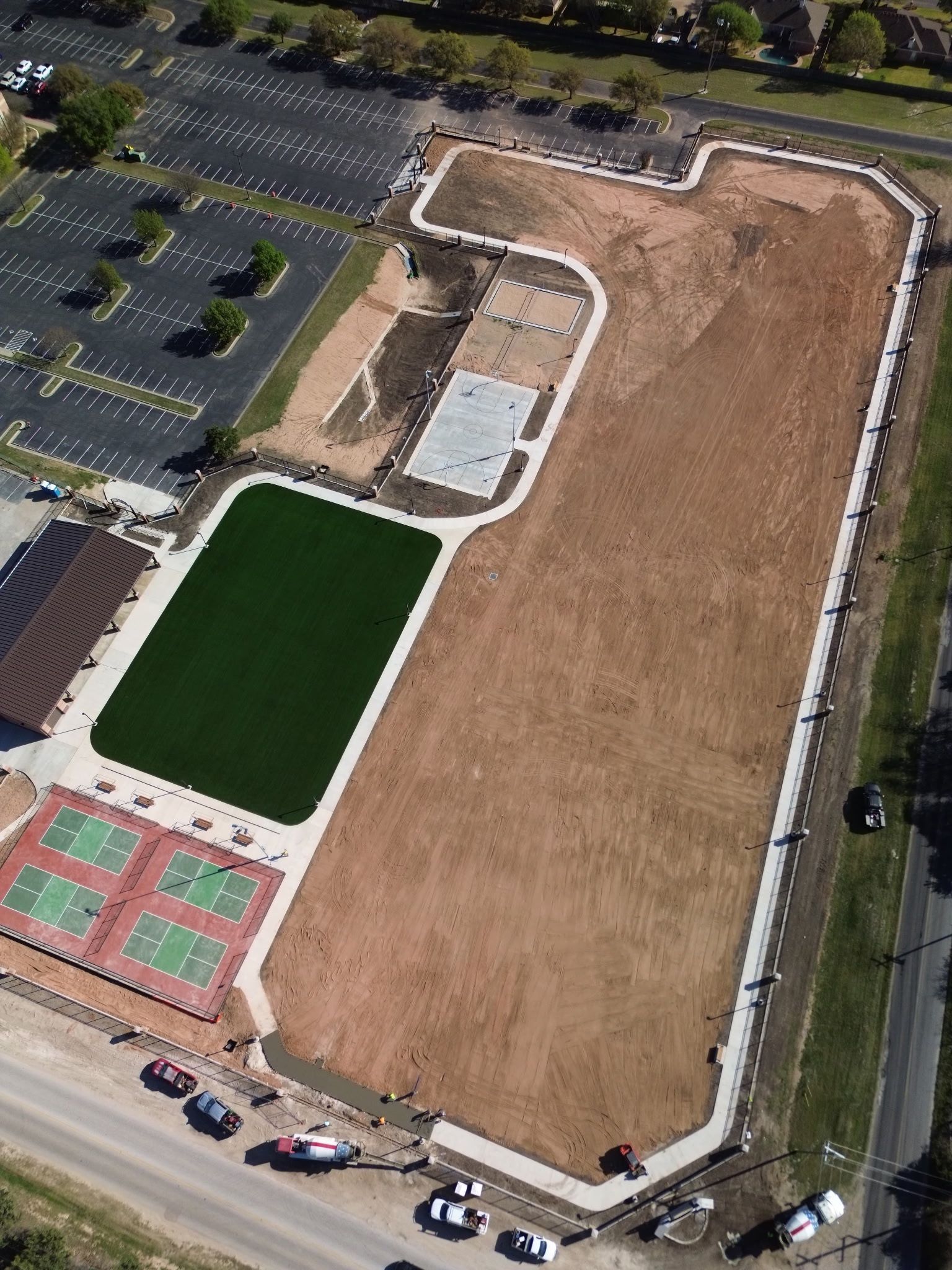 Aerial view of a large vacant dirt lot beside a parking lot, green field, and tennis courts.