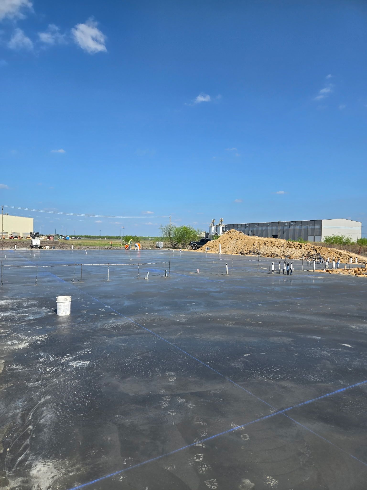 Empty paved lot under blue sky, with a few people and industrial buildings in the distance.