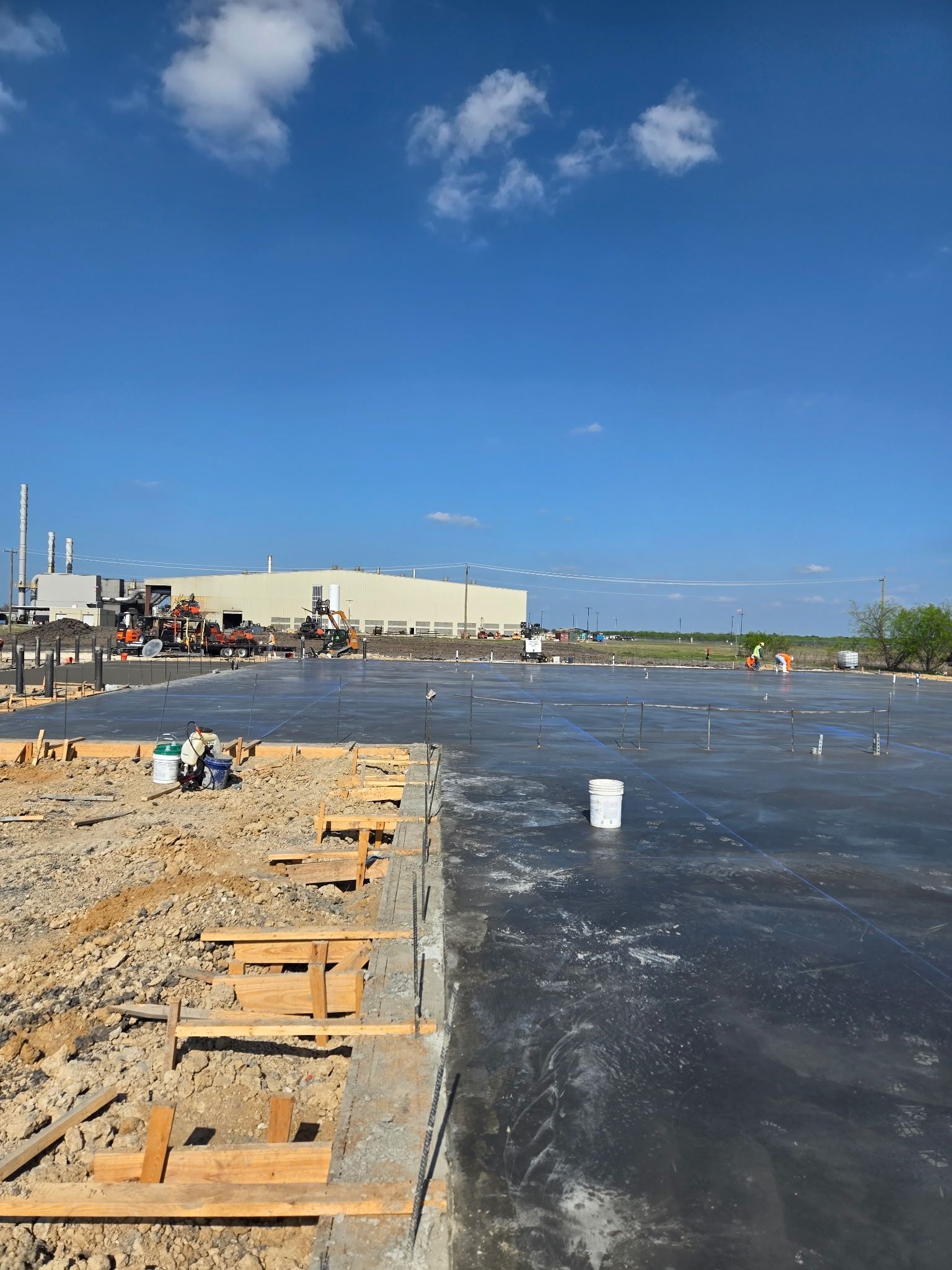 Beach shoreline beside a rippling river under a blue sky, with people and buildings in the distance.