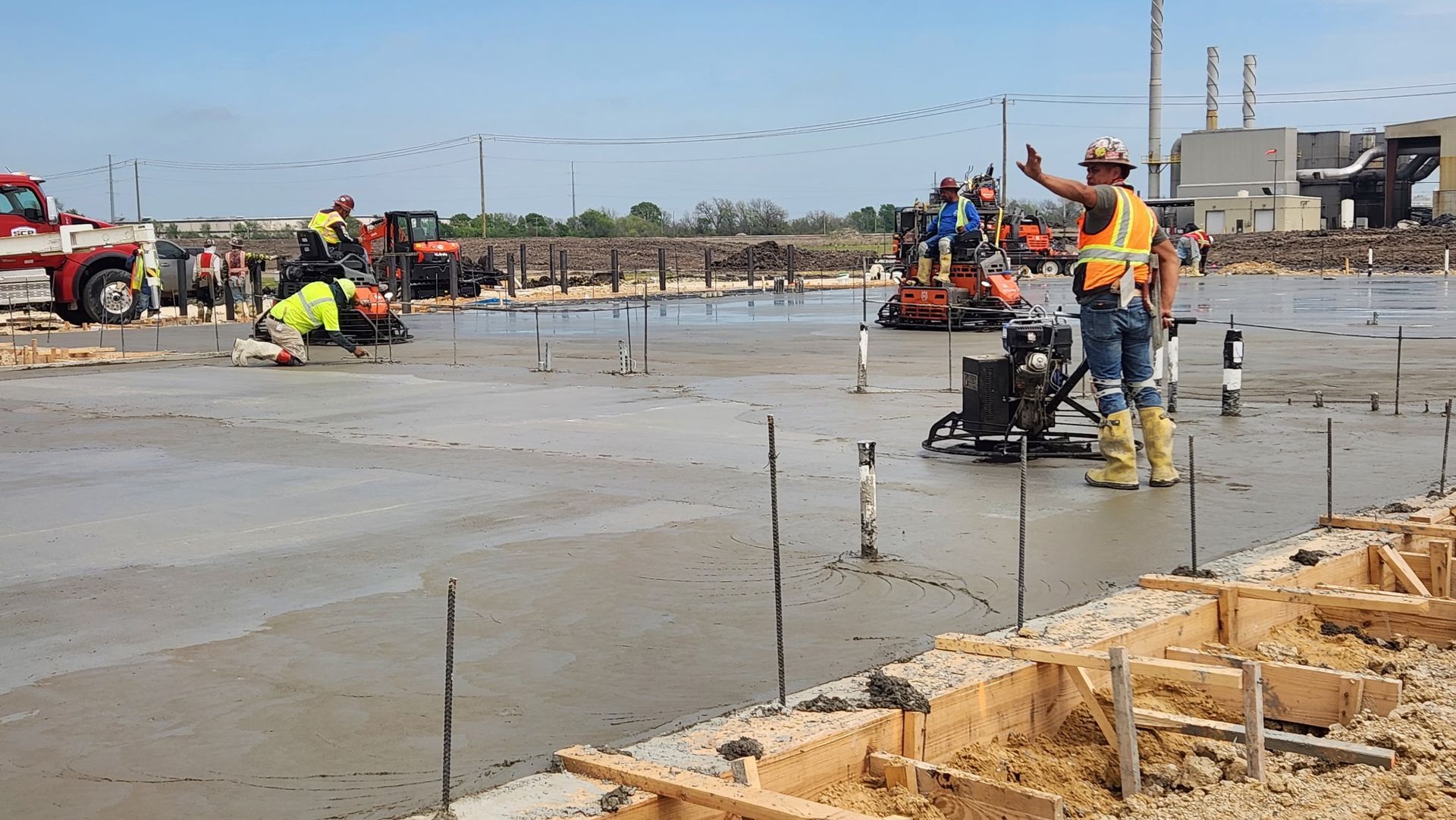 Workers smoothing wet concrete at a construction site, with tools, rebar, and a flatbed truck in the background