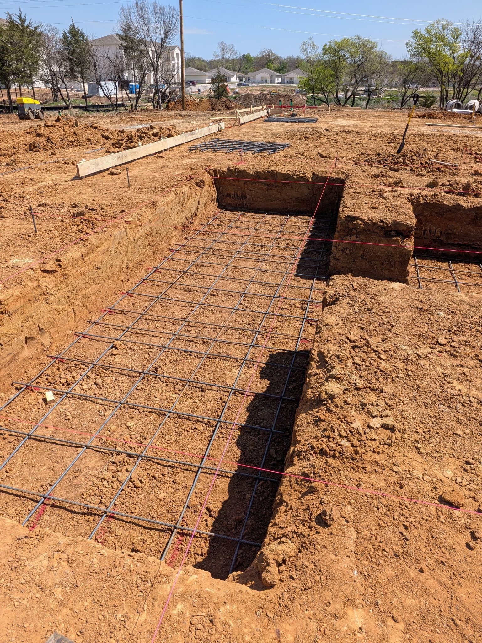 Rebar foundation slab under construction on a sandy lot, with trenches and a bright blue sky.