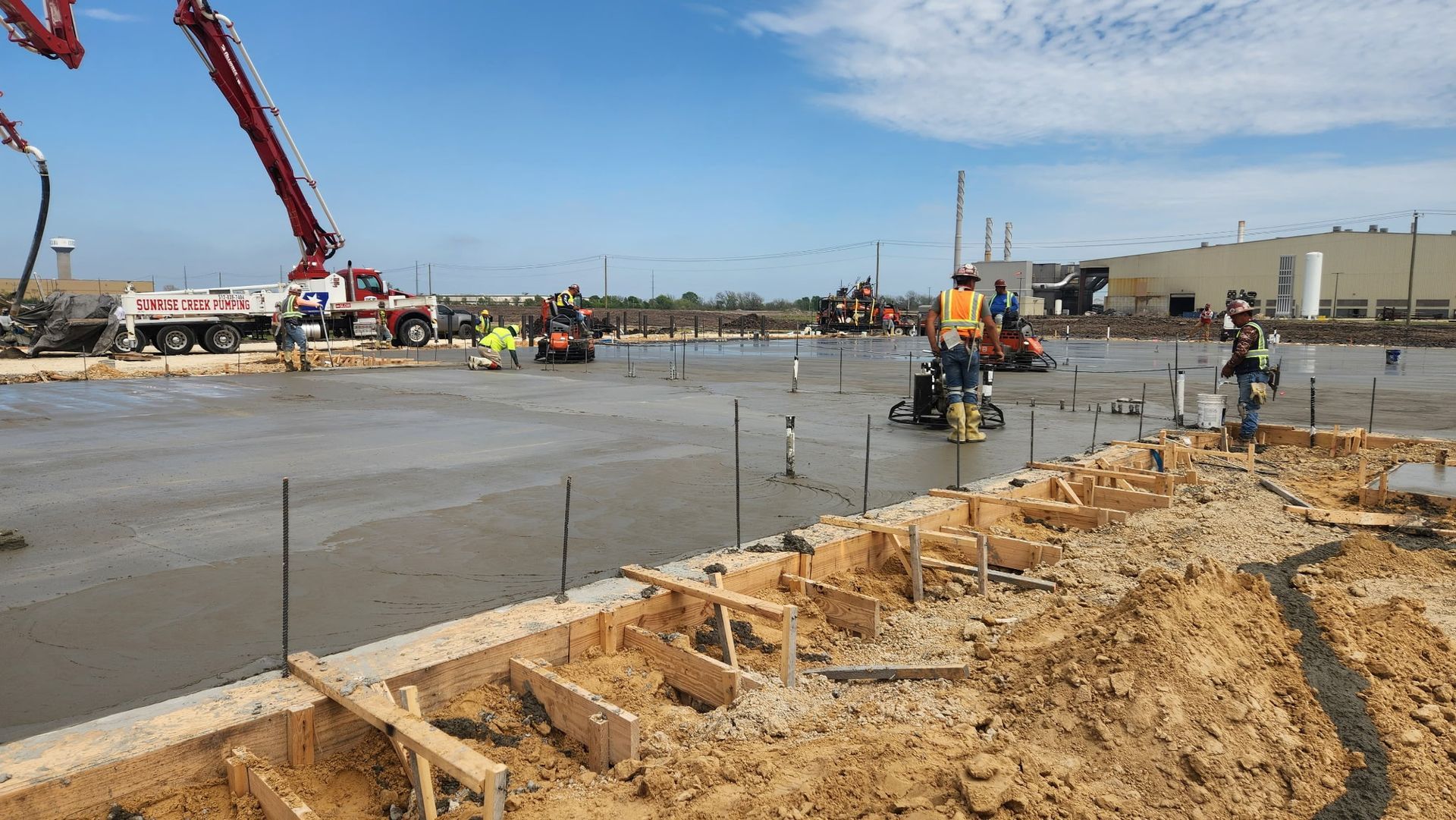 Construction site with concrete slab being poured, workers, cranes, and dirt piles under a clear sky
