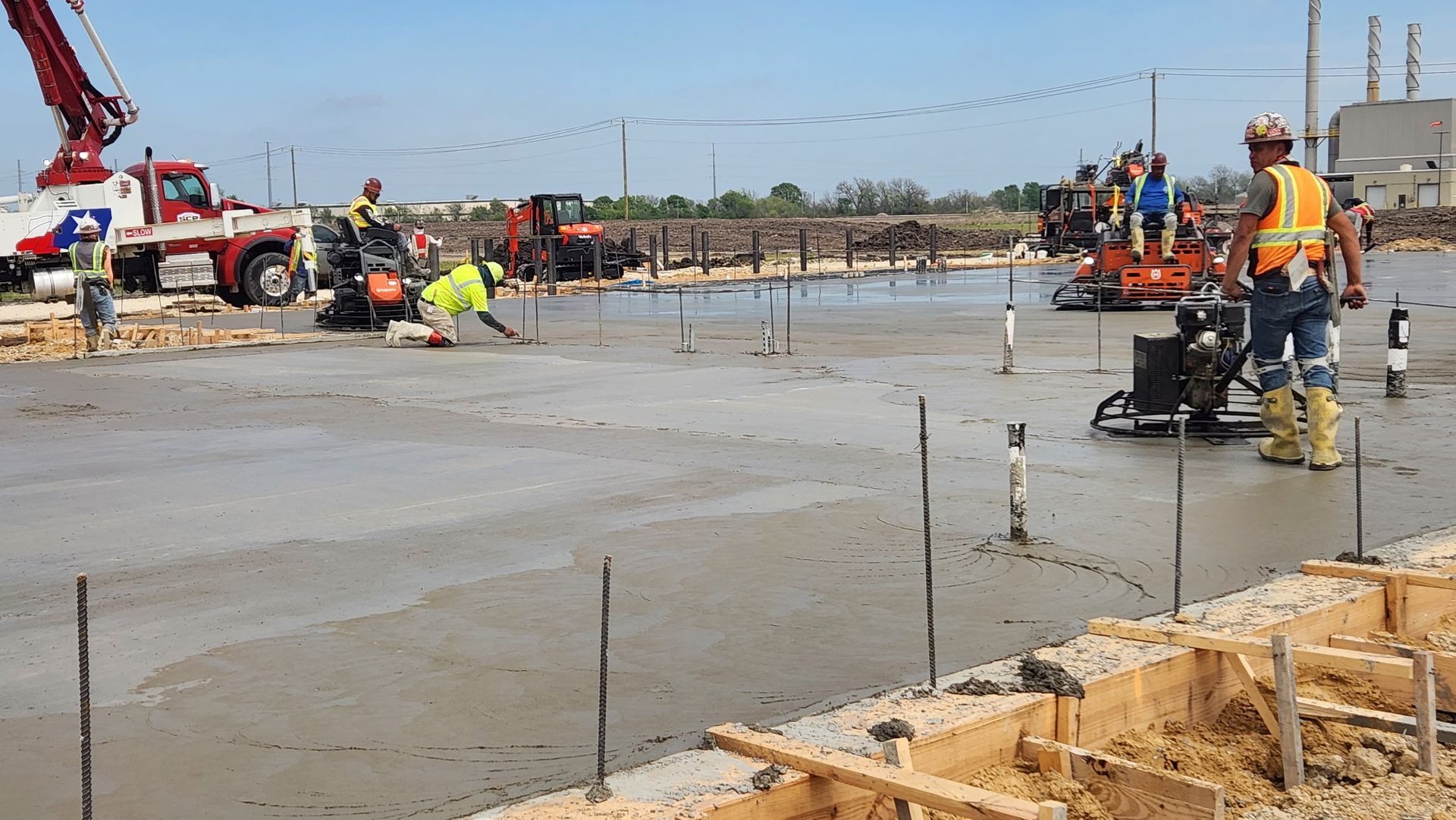 Construction workers finishing a concrete slab, with a power trowel and equipment on a wet worksite.