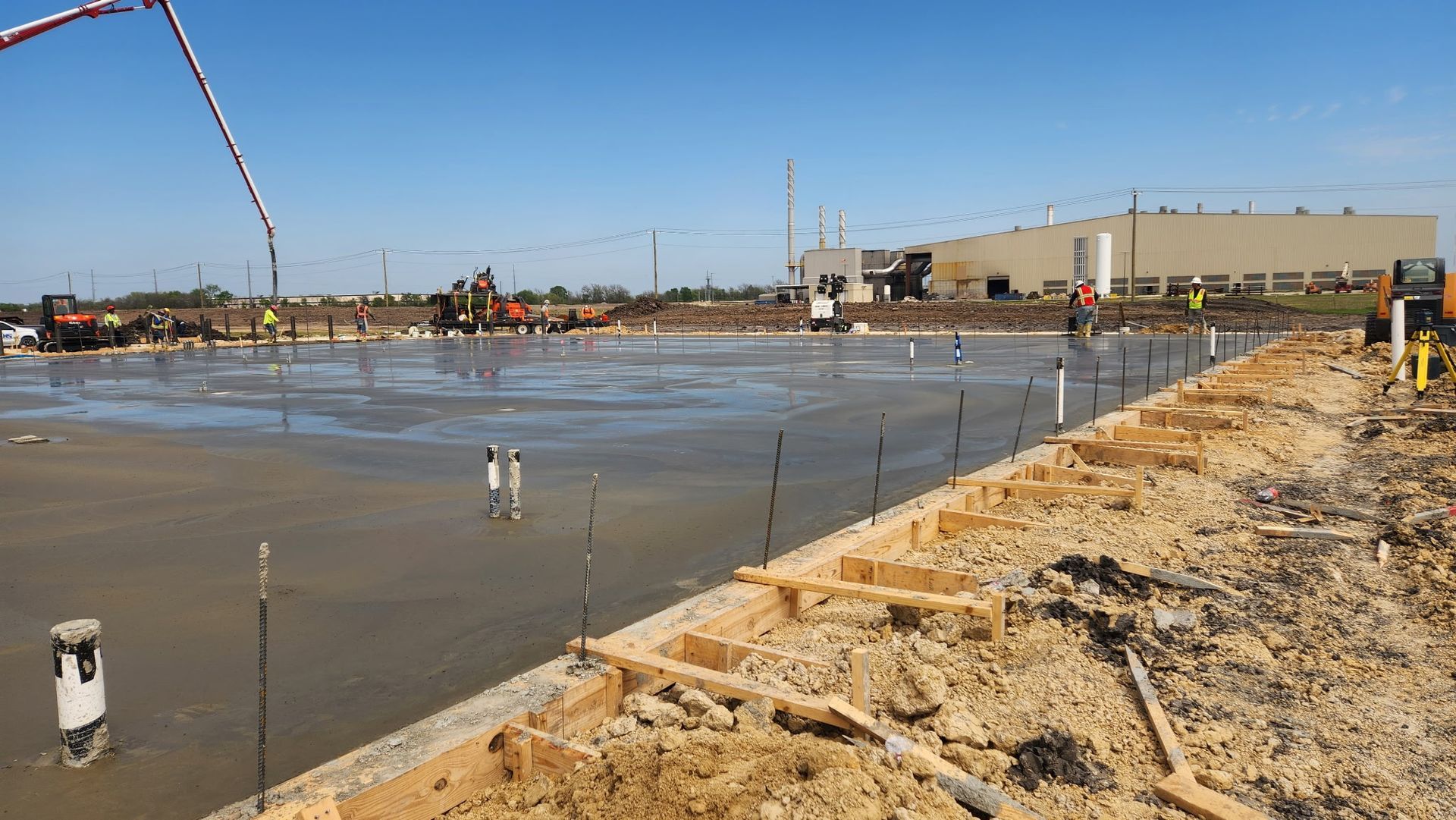 Construction site with wet concrete slab, muddy ground, and a long boom pump under a clear blue sky