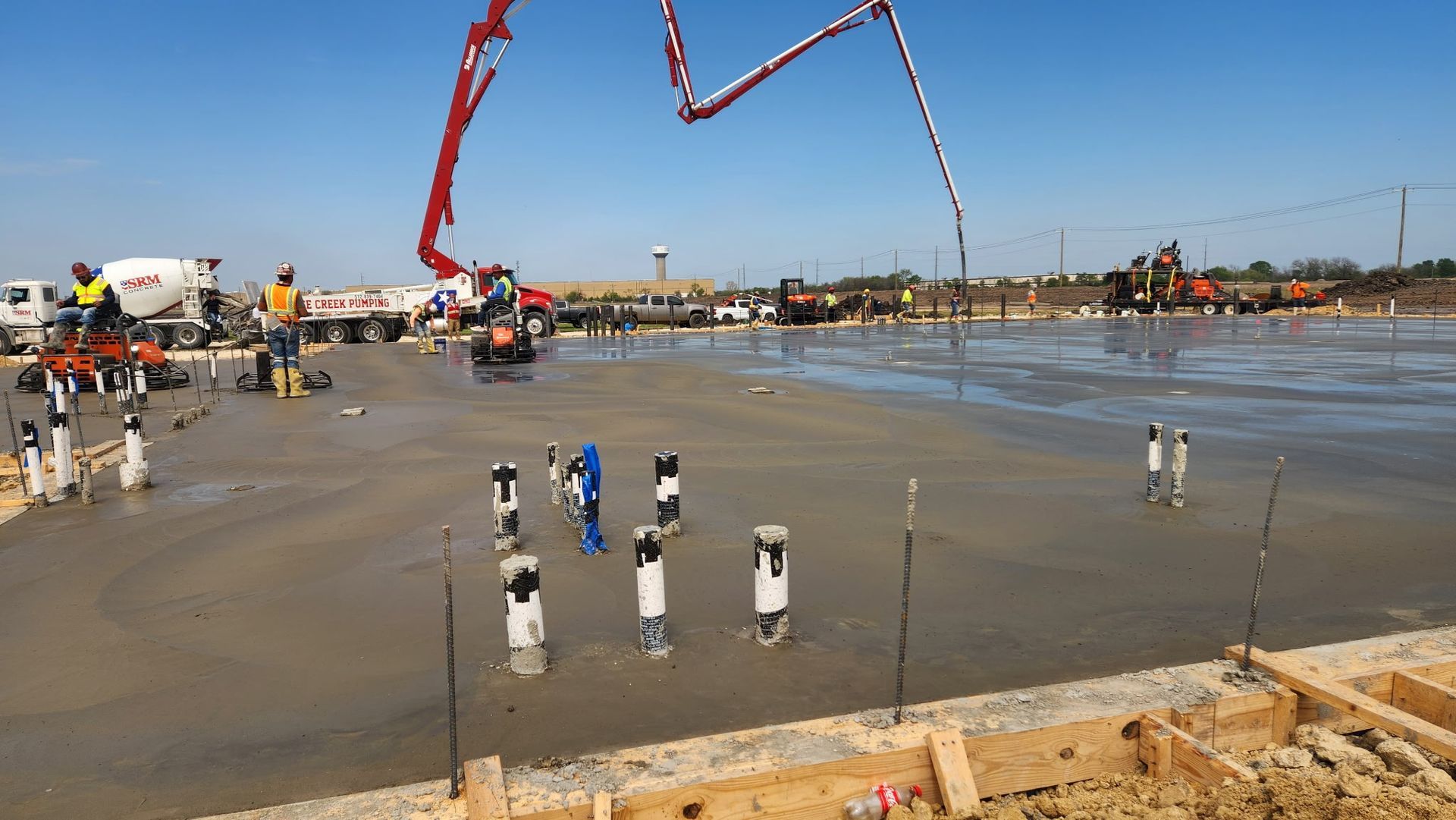 Workers at a concrete construction site with red boom pumps and wet ground near water.