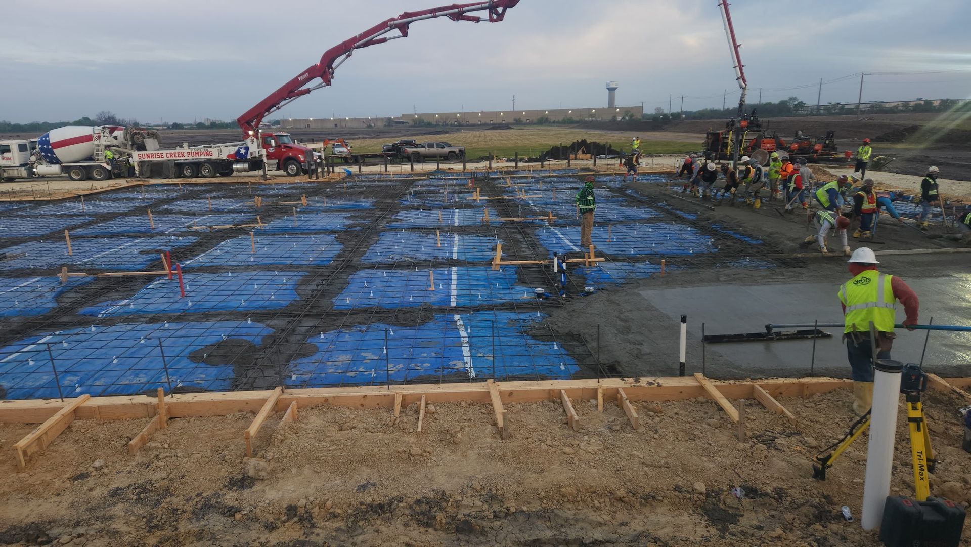 Construction site with large blue-covered foundation, concrete being poured, and a crane in the background
