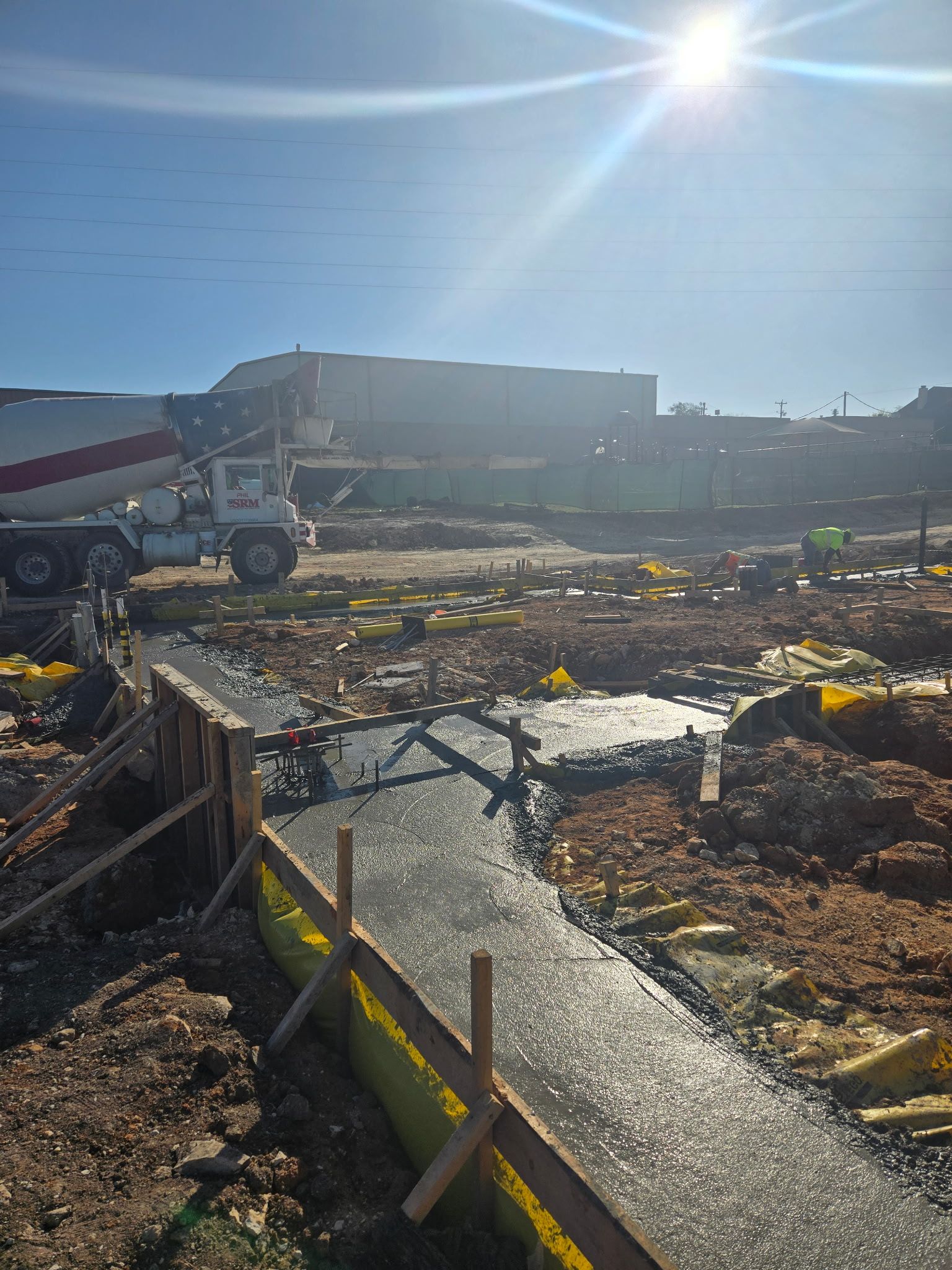 Construction site with wet concrete walkway, excavator, and bright sun over a muddy area