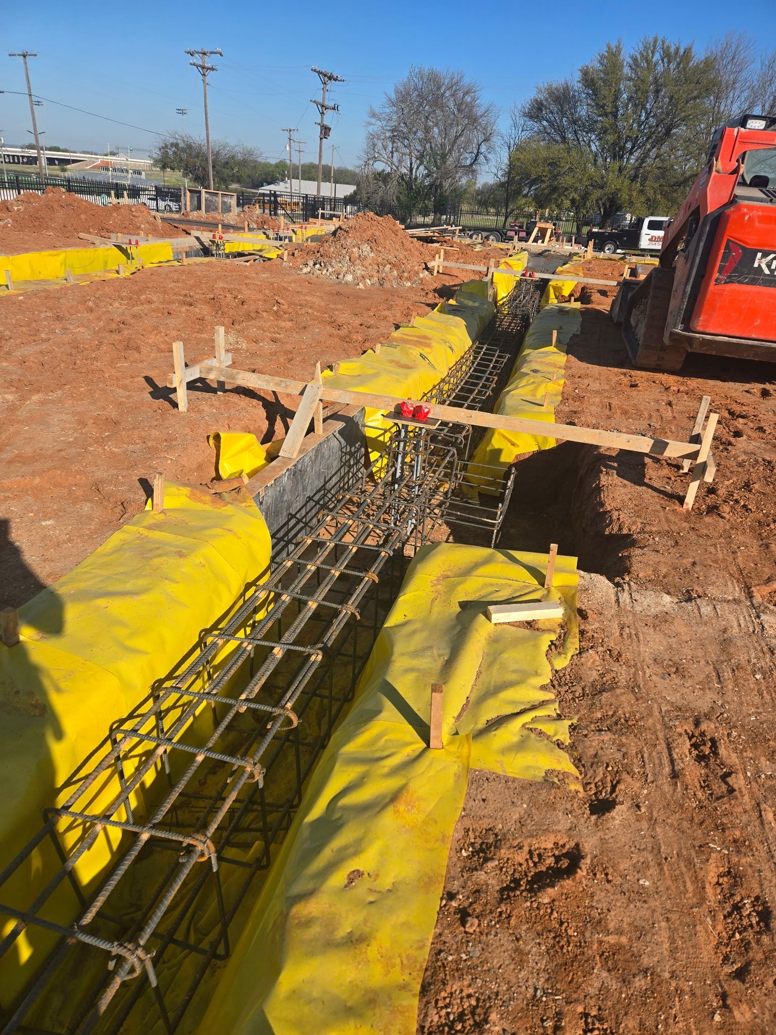 Construction site with yellow-covered rebar trench and an excavator on the right.