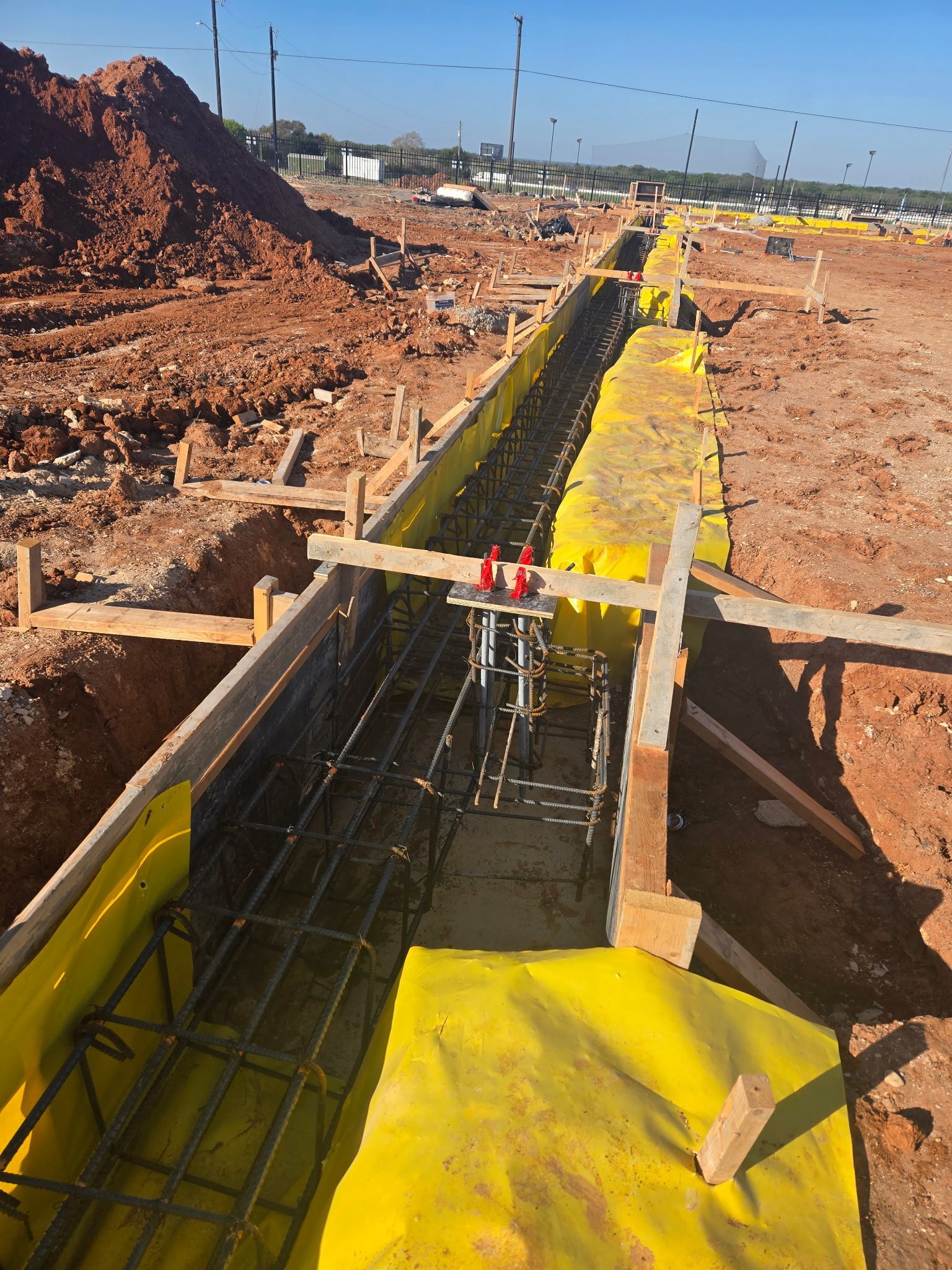 Construction site trench with yellow safety lining and rebar beside piles of dirt under a clear sky