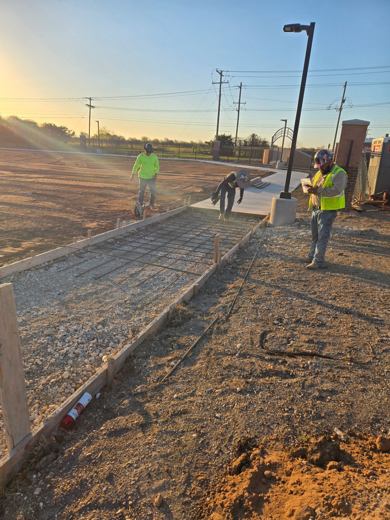 Workers in neon vests on a gravel roadside at sunset, near utility poles and a concrete drainage channel