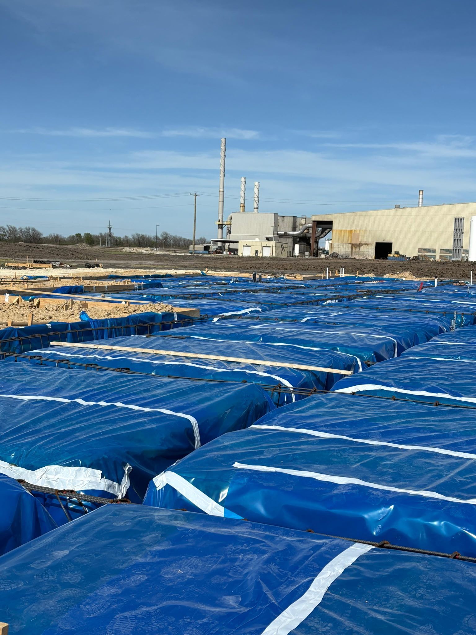Rows of blue tarps covering ground near an industrial site under a clear sky.