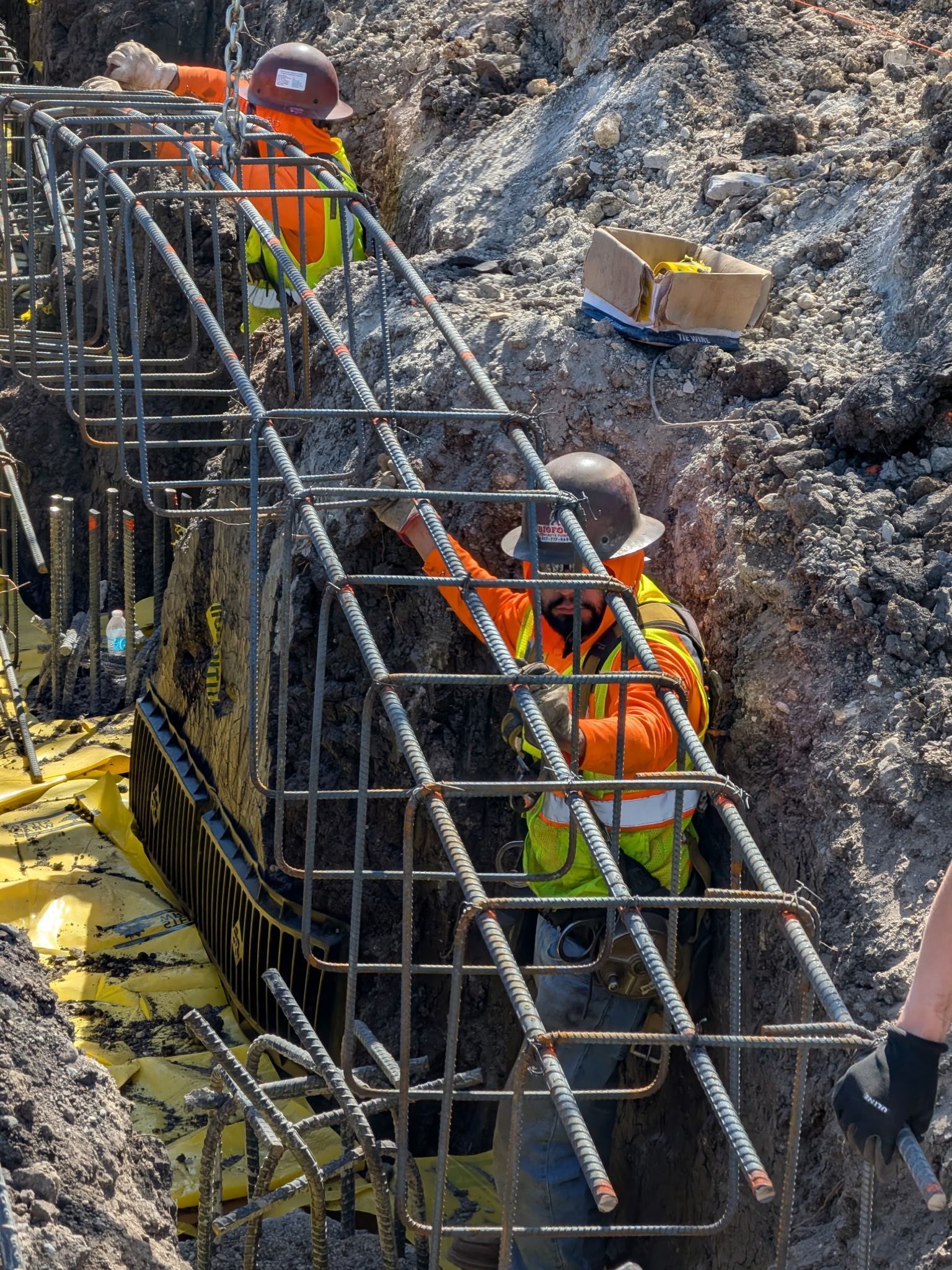 Workers in orange safety gear inspect rebar at a construction site beside a rocky trench