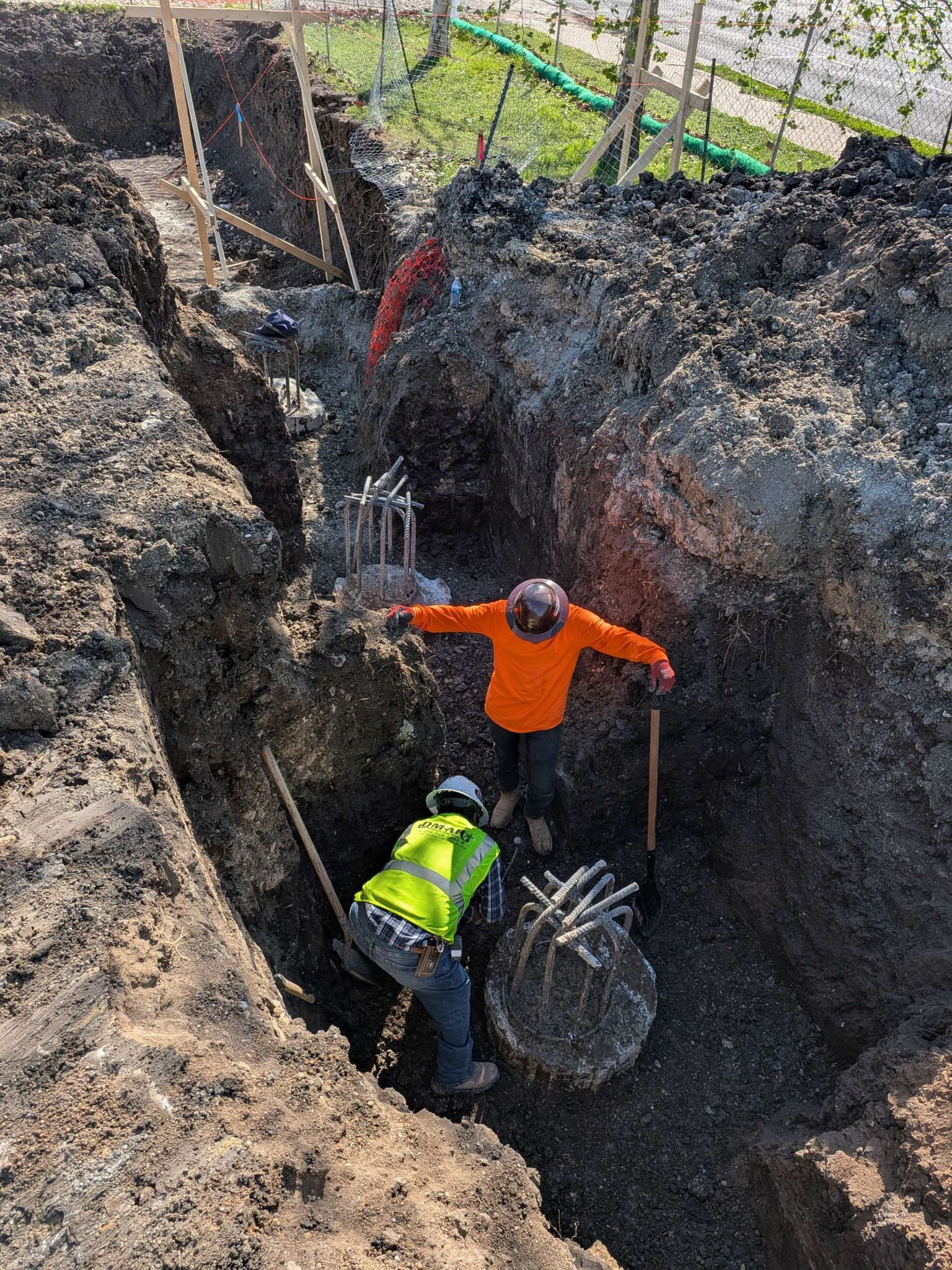 Two workers in a deep trench with a large pipe, one in orange and one in neon yellow, digging in muddy soil