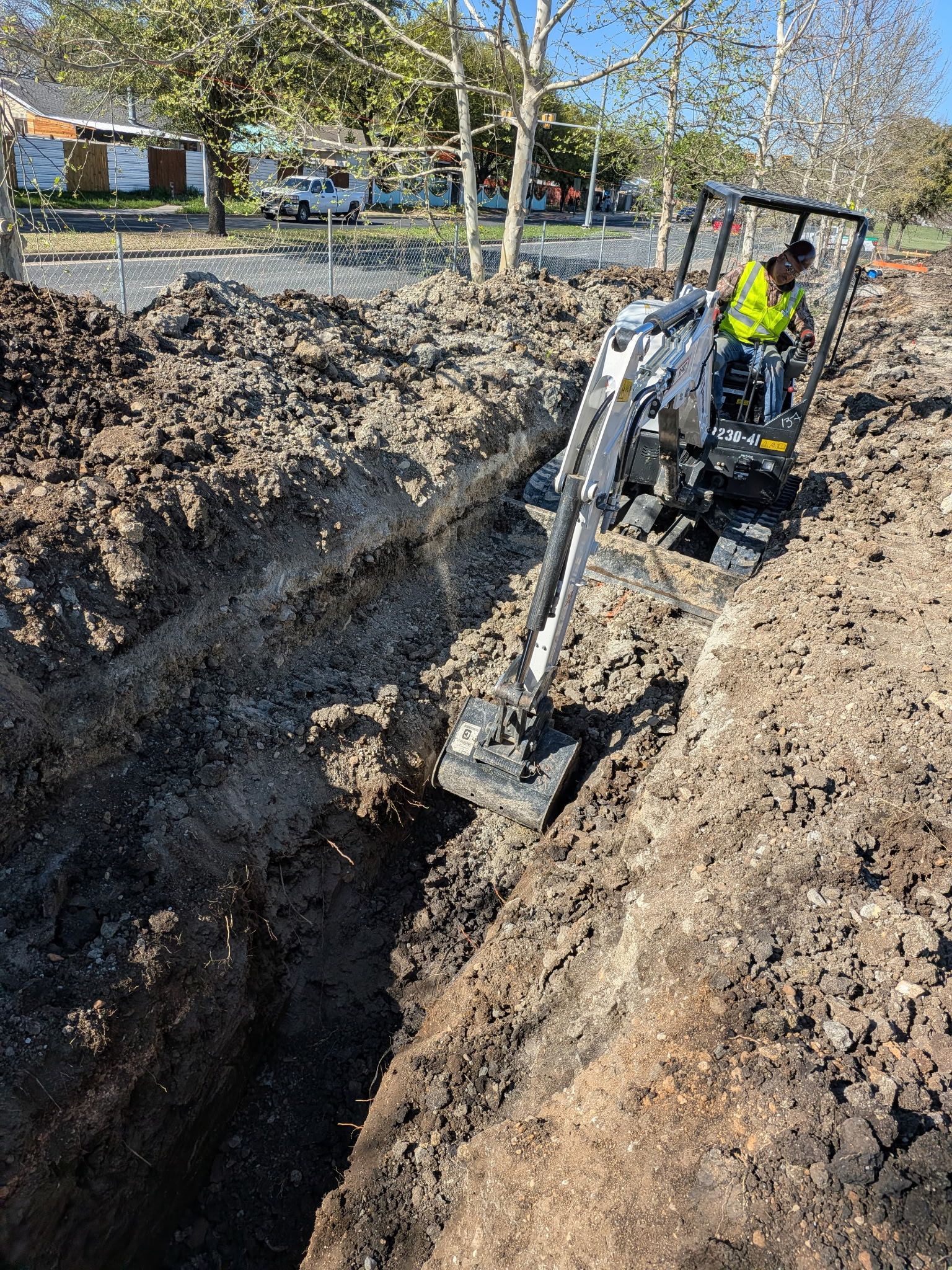 Excavator digging a trench in a muddy yard, with a worker in a yellow jacket nearby