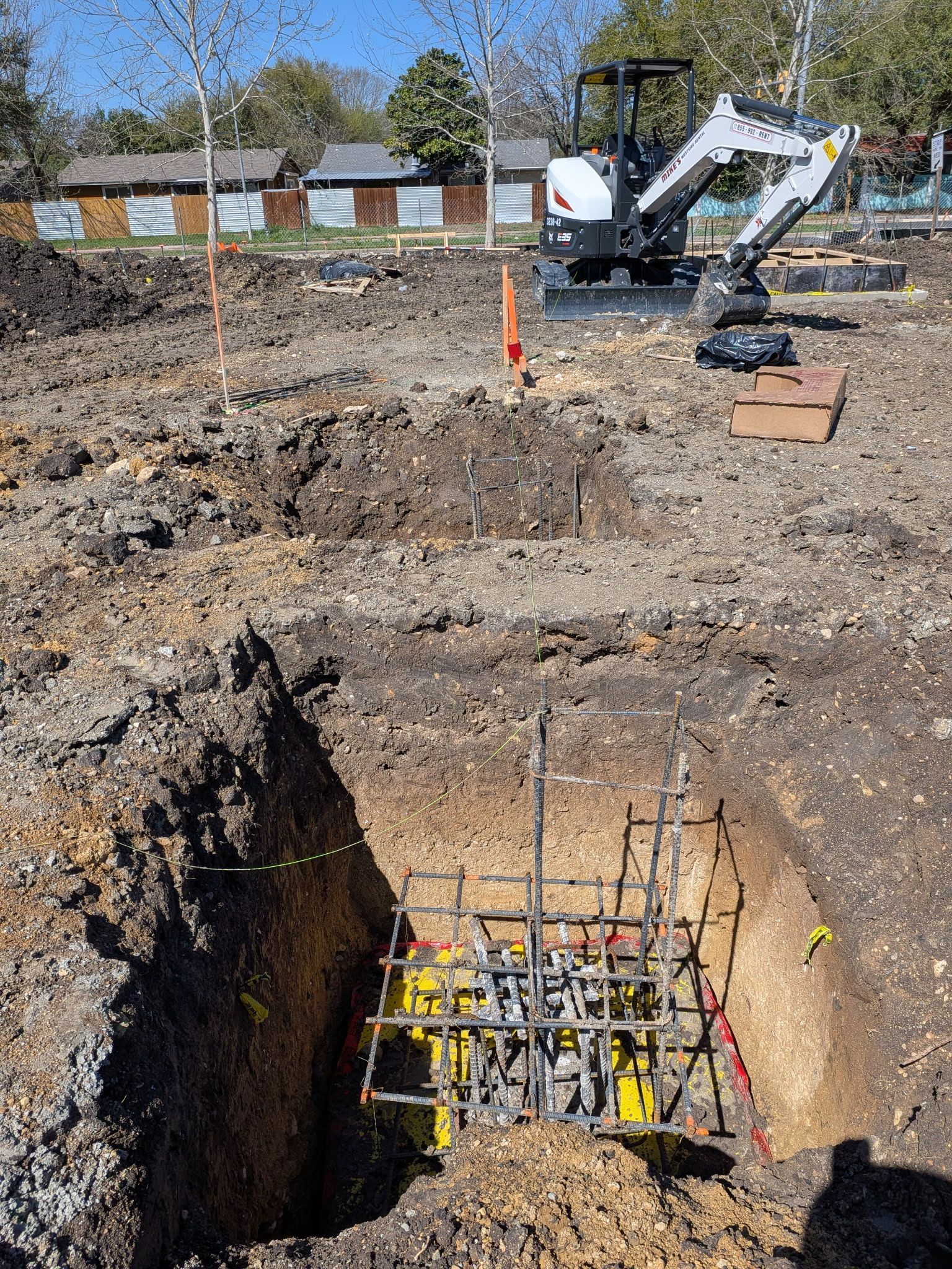 Construction site with excavated hole, rebar foundation, and a skid steer in the background
