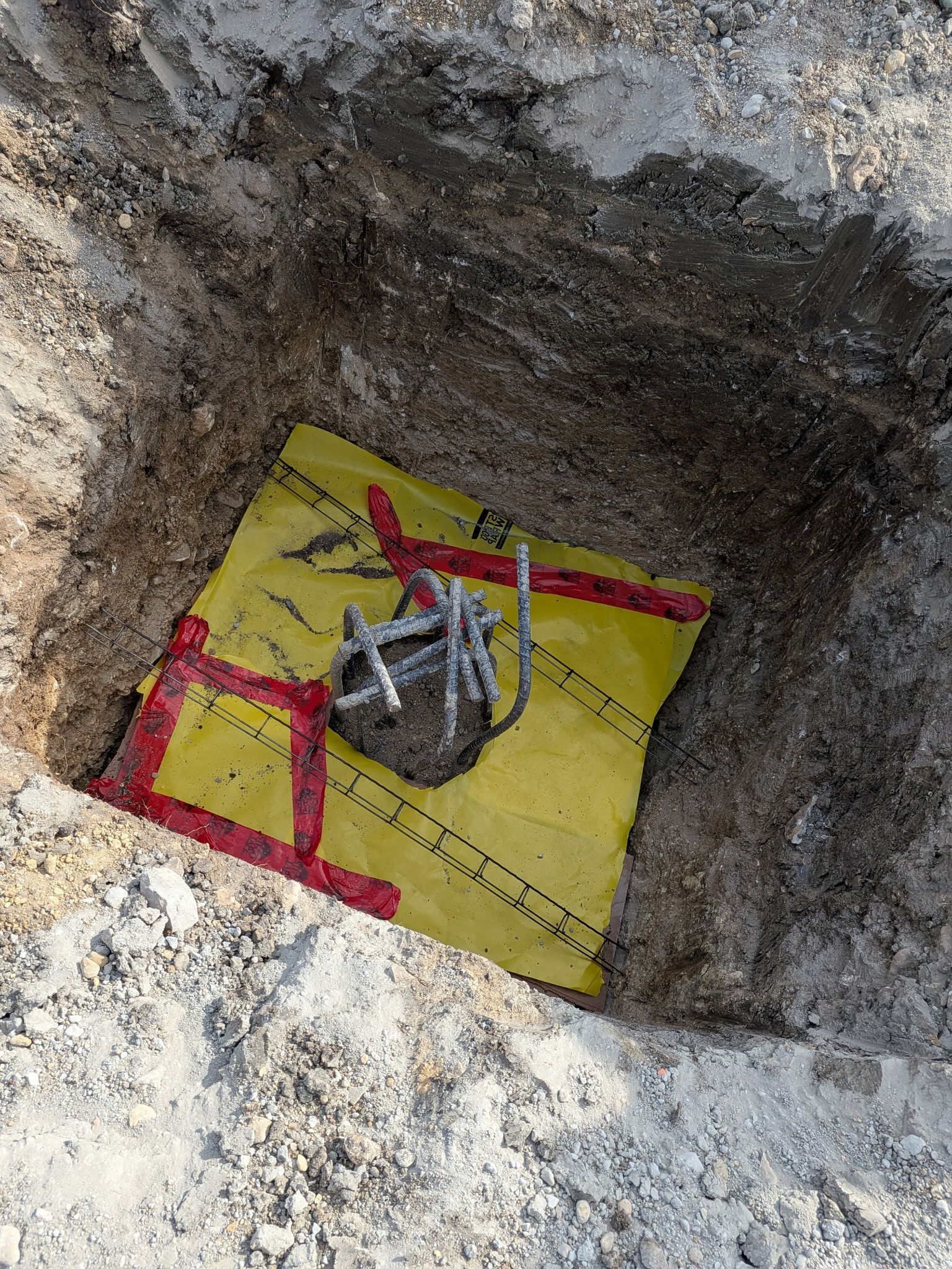 Open excavation pit with a yellow board, red markings, and a metal pipe or fitting in the center.