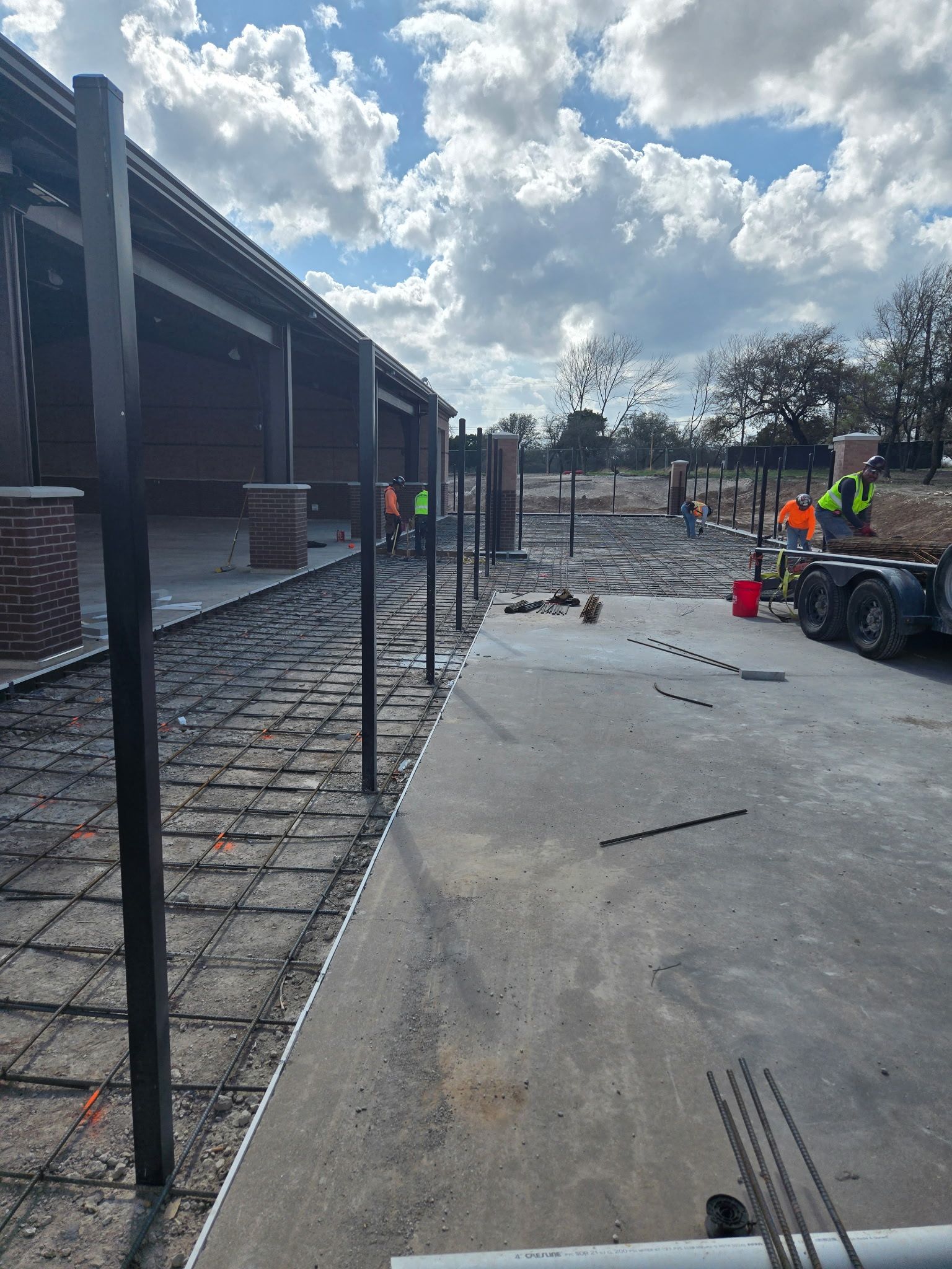 Construction site with workers, red buckets, and metal posts beside a building under a cloudy sky