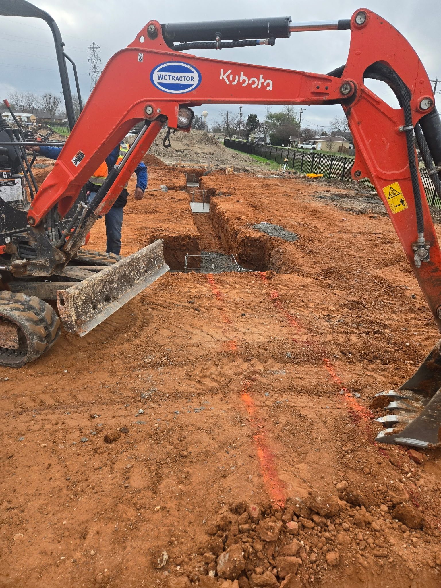 Red excavator leveling a dirt lot at a construction site, with rubble and muddy ground.