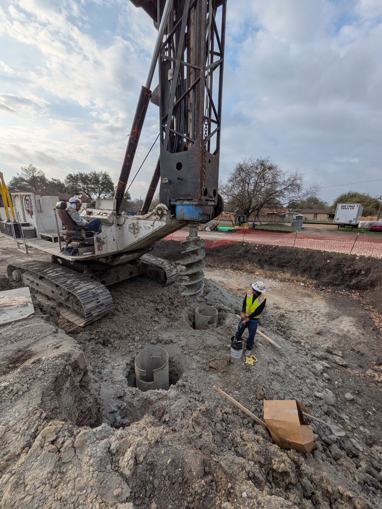 Construction site with drilling rig excavating a deep trench and a worker in a yellow vest nearby