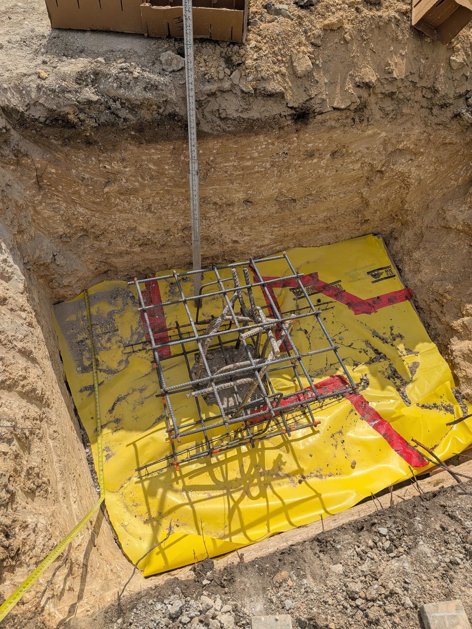 Construction footing excavation with rebar cage over a yellow waterproofing membrane in a dirt pit