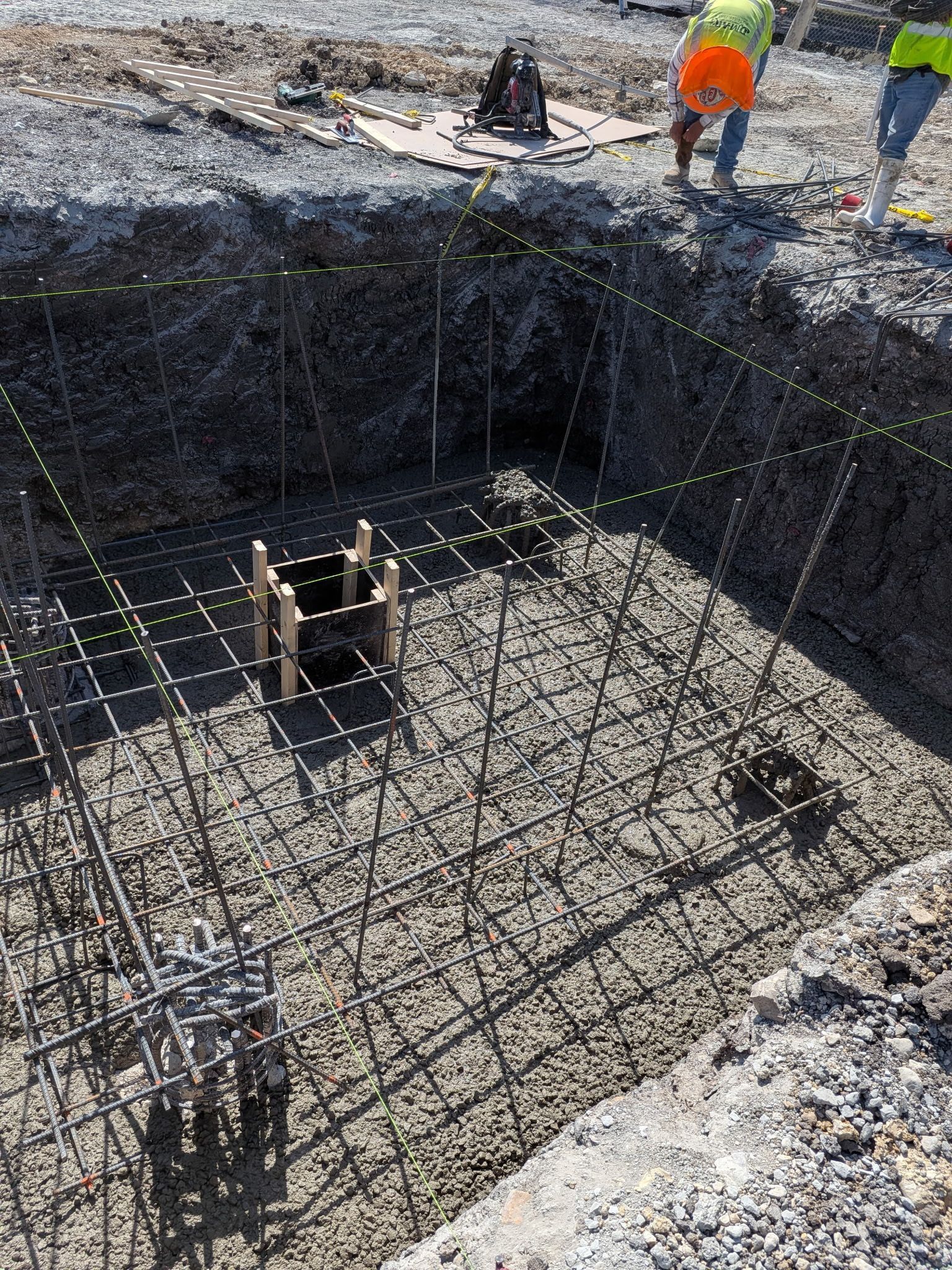 Large excavated hole with rebar foundation grid and workers in orange safety gear above ground
