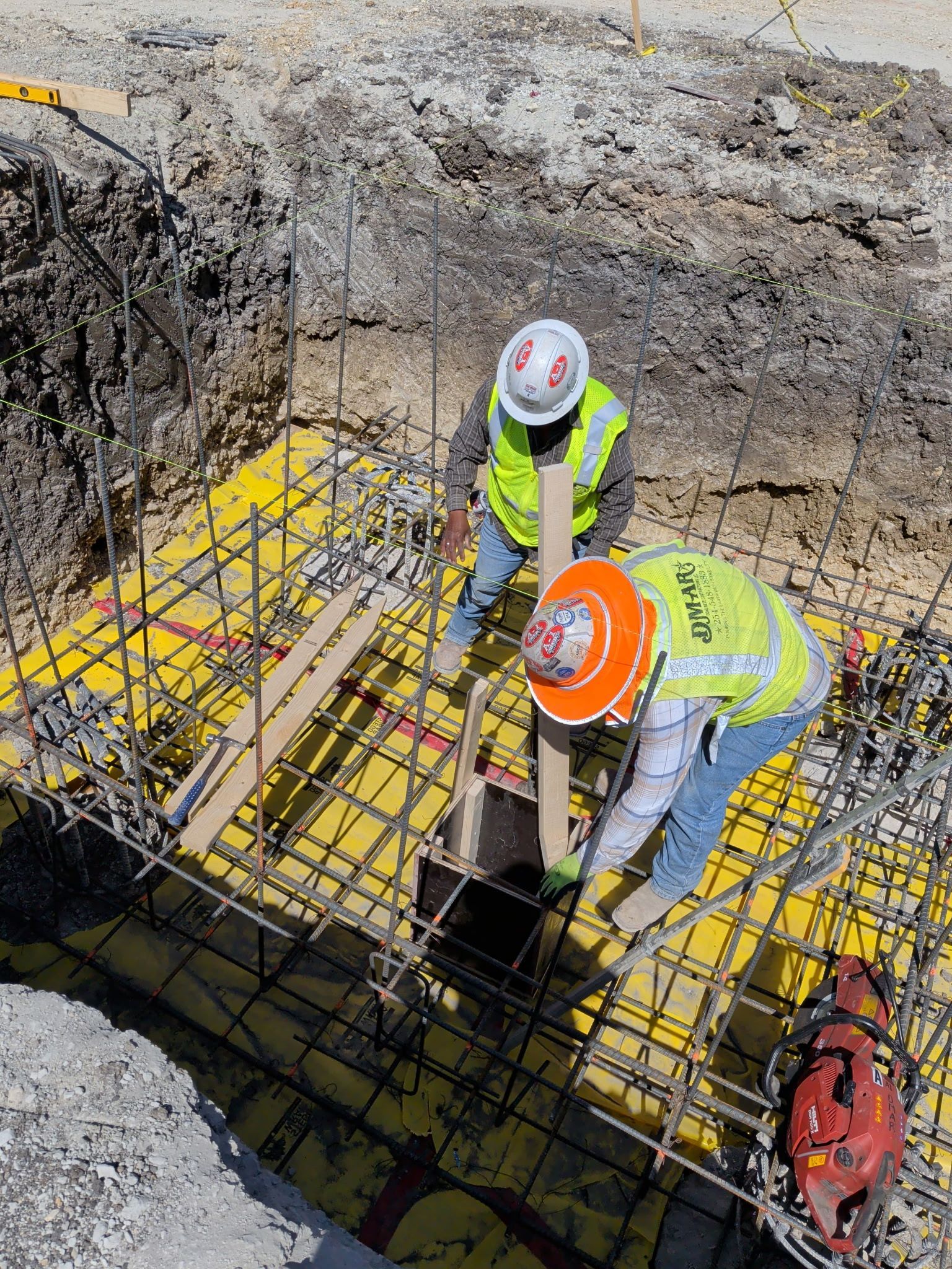 Two workers in hard hats inspect rebar at a concrete foundation excavation site.