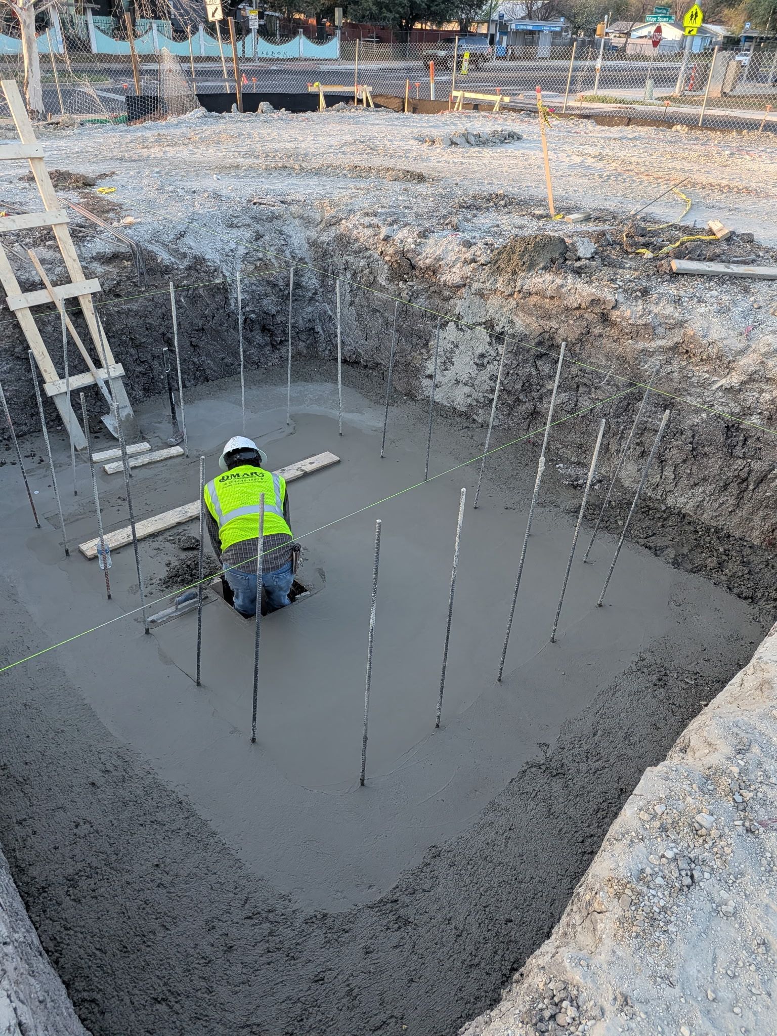 Worker in neon safety vest kneels in wet concrete at a construction site with rebar and a ladder.
